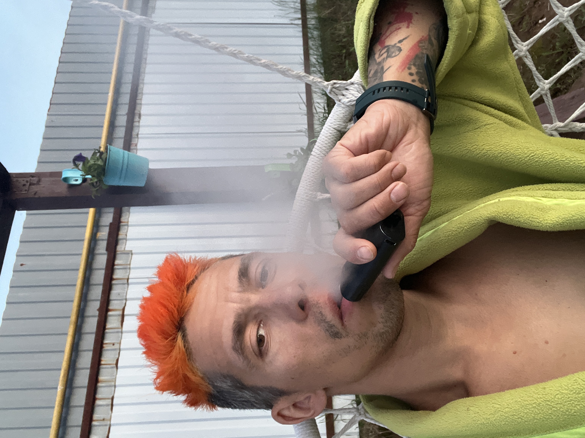 This close-up, vertically oriented image captures a person outdoors in Chaikovsky, Russia, relaxing and exhaling vapor from a device. The subject, a person with distinct orange-dyed hair and light stubble, looks directly at the camera with a neutral expression. They are casually draped in a light green, fuzzy blanket or robe, revealing their bare chest and a tattooed left arm adorned with a black smartwatch. White smoke or vapor billows from their mouth as they hold a black vaping device.
The background elements suggest an outdoor setting. Visible are thick white ropes, indicating the person is likely reclining in a hammock. A corrugated metal wall or fence provides the backdrop, with a bright, clear sky visible above. To the left, a light blue hanging flower pot containing a small plant is attached to a wooden beam. A slender yellow pole and a brown beam are also visible vertically against the corrugated siding. The scene conveys a moment of casual relaxation. No visible text is present in the image.