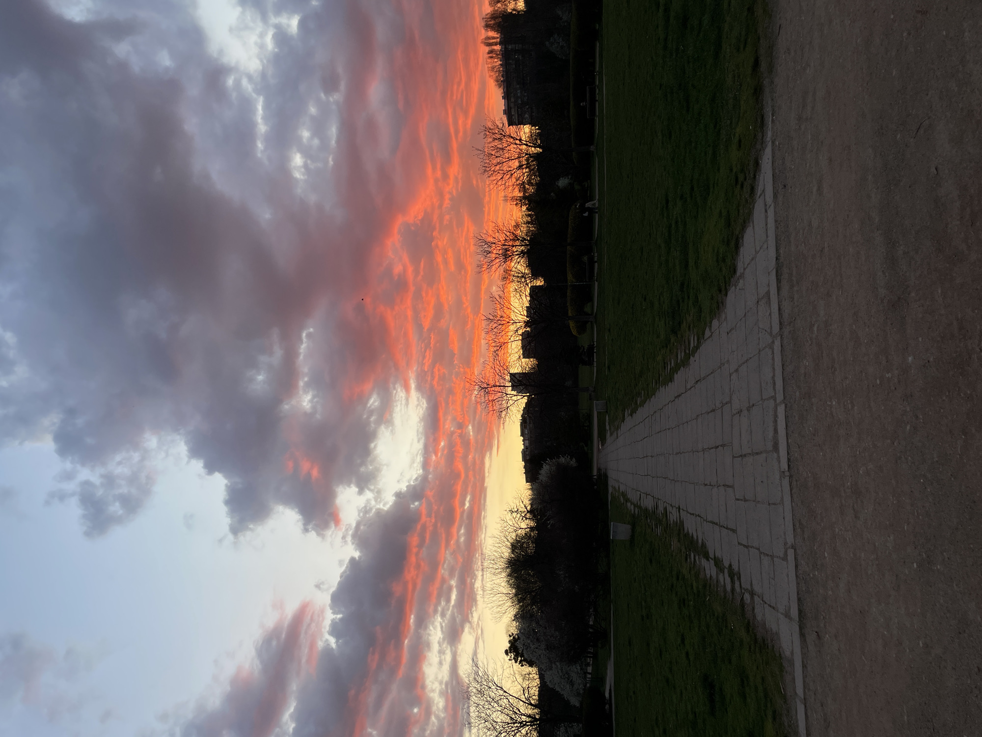 The image depicts a park setting in Sofia, Bulgaria, at dusk or dawn. A stone-paved pathway extends into the distance, bordered by grass on one side and an unpaved, dirt path on the other. Trees and buildings are visible in the background. The sky is filled with clouds colored in hues of orange, pink, and gray, reflecting the light of the setting or rising sun.