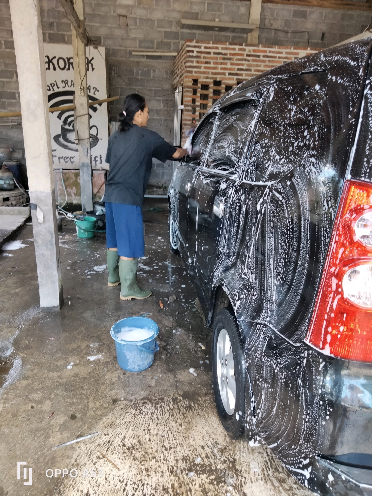A person is washing a black car with soap suds at an outdoor car wash facility in Ngadirenggo, Indonesia. The person is wearing a black shirt, blue shorts, and green rubber boots. They are standing next to the car, holding a sponge and reaching towards the car's window. A blue bucket filled with soapy water is on the ground in front of the person. The car wash area appears to be a concrete floor with puddles of water and soap. In the background, there is a brick wall with some signs and exposed pipes. The overall environment suggests a functional, no-frills car wash service.