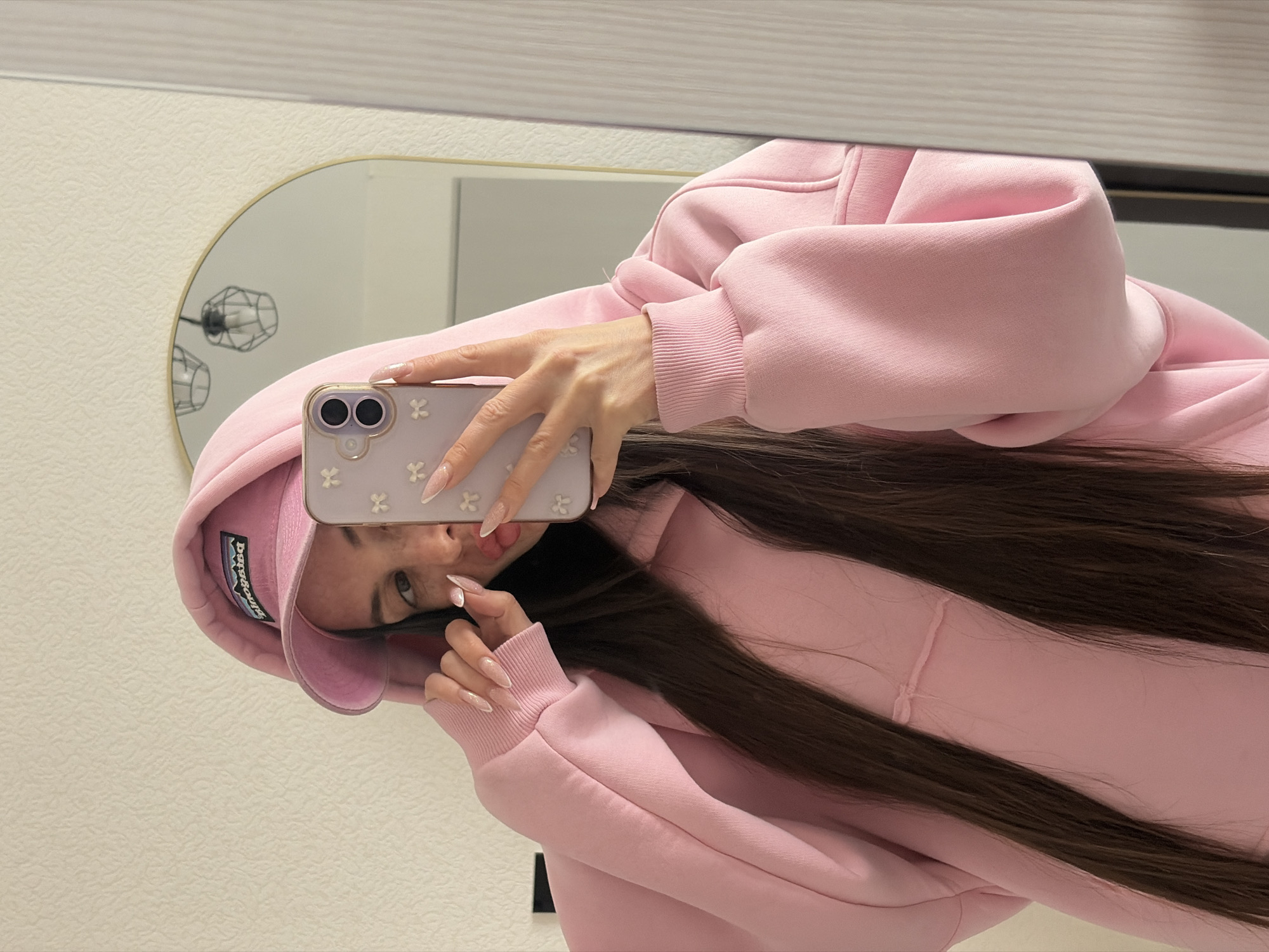An indoor mirror selfie, rotated 90 degrees clockwise, shows a person (likely female) wearing an oversized pink hoodie and a matching pink baseball cap. The individual has long, straight, dark brown hair that cascades down. They are holding a light-colored smartphone with a case adorned with white four-leaf clover or floral patterns, revealing two camera lenses on the back. The person's nails are long, manicured, and a light, possibly nude or pale pink shade with a subtle glitter finish. One eye is visible, looking directly at the camera, and their mouth is slightly open, with a glimpse of the tongue, conveying a casual or playful expression. The baseball cap clearly displays the "Patagonia" logo.
The selfie is taken in an oval-shaped mirror with a thin gold or brass frame. Reflected in the mirror is a geometric black wire light fixture and a light-colored wall. The actual wall visible in the foreground is also light-colored and has a subtle textured pattern. The scene depicts a casual moment inside a room, located in Saint Petersburg, Russia.