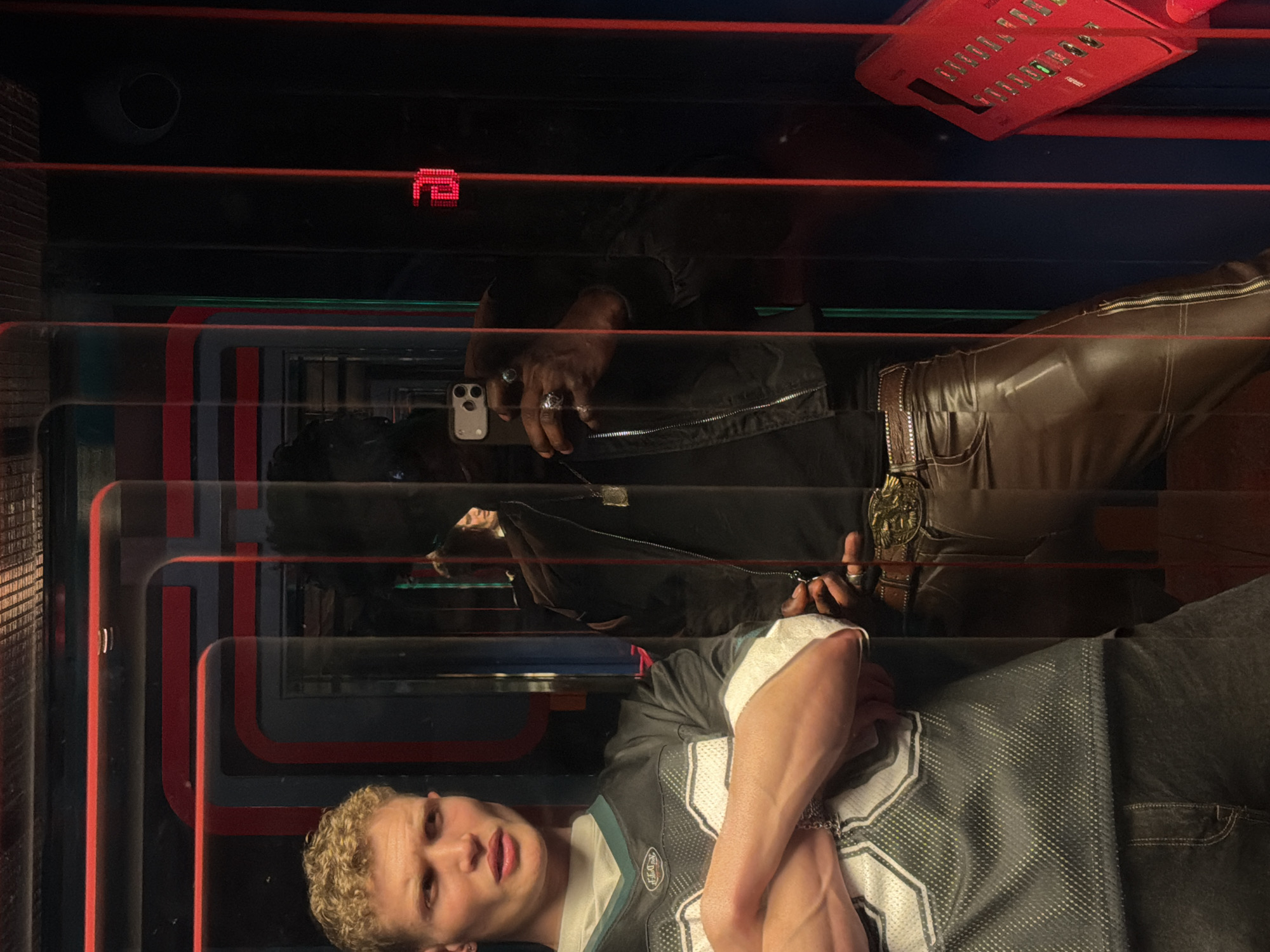 The image shows a reflection of three people in what appears to be a retro-themed setting. A young man with short, light-colored hair wearing an Eagles jersey is at the bottom of the frame, with his arms crossed. Above him is a reflection of a dark-skinned person with a black jacket, brown leather pants, and a large belt buckle. They are holding a smartphone with multiple rings on their fingers. At the top, the reflection shows a black-clad individual holding another phone.
The setting features red, black, and grey rectangular patterns. Elements like a red keyboard with buttons, and a cylindrical object, possibly a speaker, are visible. The overall impression is of a bar or entertainment venue with a retro, 1980s aesthetic. While the style is evocative of locations within London, United Kingdom, there is no explicit evidence to pinpoint the scene's location to the city.