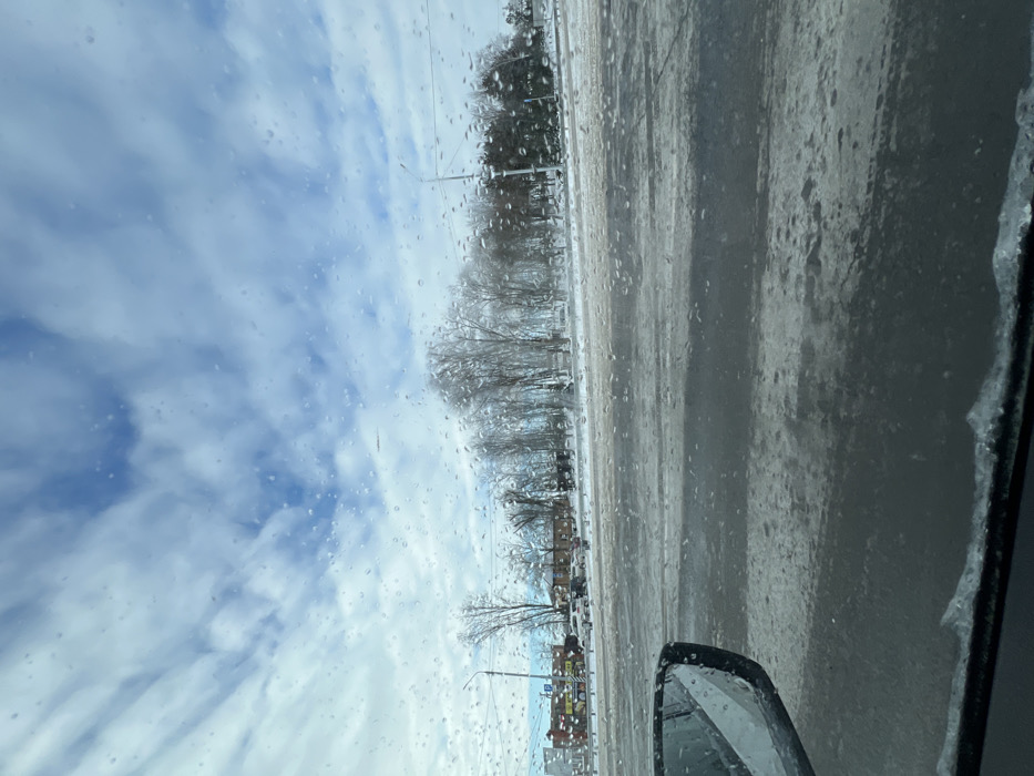 The image depicts a winter street scene in Sambek, Russia, viewed from inside a vehicle. The foreground shows the lower-left section of a vehicle's windshield and a side mirror, both partially obscured by water droplets and ice accumulation along the bottom edge of the windshield. The midground features a multi-lane asphalt road, partially covered with patches of snow and ice, indicating wintry conditions. To the left of the road, a commercial area is visible with several low-rise buildings, some displaying colored signage, and a parking lot containing multiple parked automobiles. A streetlight post stands on the left. To the right of the road and extending towards the center, a dense line of mostly bare deciduous trees and some evergreens suggests a park or wooded area. Another streetlight post and overhead power lines are visible on the right. The background consists of a sky with scattered white and light-grey clouds against a blue backdrop, also viewed through the droplet-covered windshield. No individuals are visible.