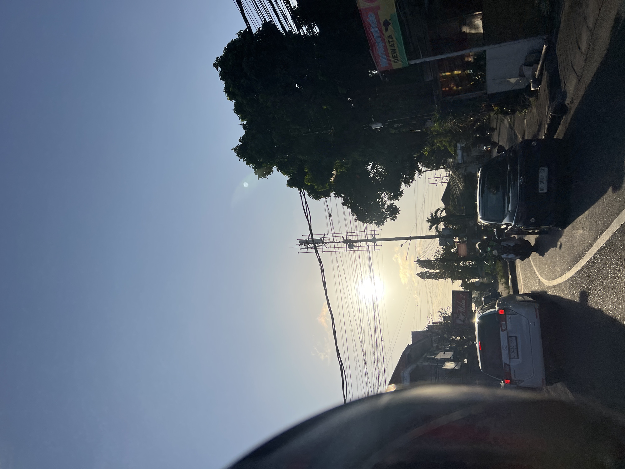 The image captures a street scene in Denpasar, Indonesia, during what appears to be late afternoon or early evening, judging by the low angle of the sun casting long shadows. The sky is a clear, pale blue, with a few wispy clouds present. The sun itself is visible, a bright orb partially obscured by a communication tower and electrical wires that crisscross the frame.
On the road, traffic is moderate. A white SUV, possibly an MPV, is in the foreground, its brake lights illuminated, indicating it is slowing or stopped. A black SUV is behind it. Several motorcycles are also present, with at least one rider visible wearing a helmet and a light-colored top.
The buildings lining the street are typical of an urban environment, with multiple stories and visible signage. One sign, partially obscured by a tree, reads "DEWATA". Lush green trees, some with dense foliage, are interspersed along the roadside and behind the buildings, suggesting a tropical climate. Electrical poles and a complex network of wires are prominent features, a common sight in Indonesian cities. The foreground on the left side of the image shows a portion of a dark-colored helmet, suggesting the image was taken from the perspective of someone riding a motorcycle. The road surface is asphalt, marked with white lines indicating lanes. The overall impression is of a busy, everyday street scene in Denpasar.