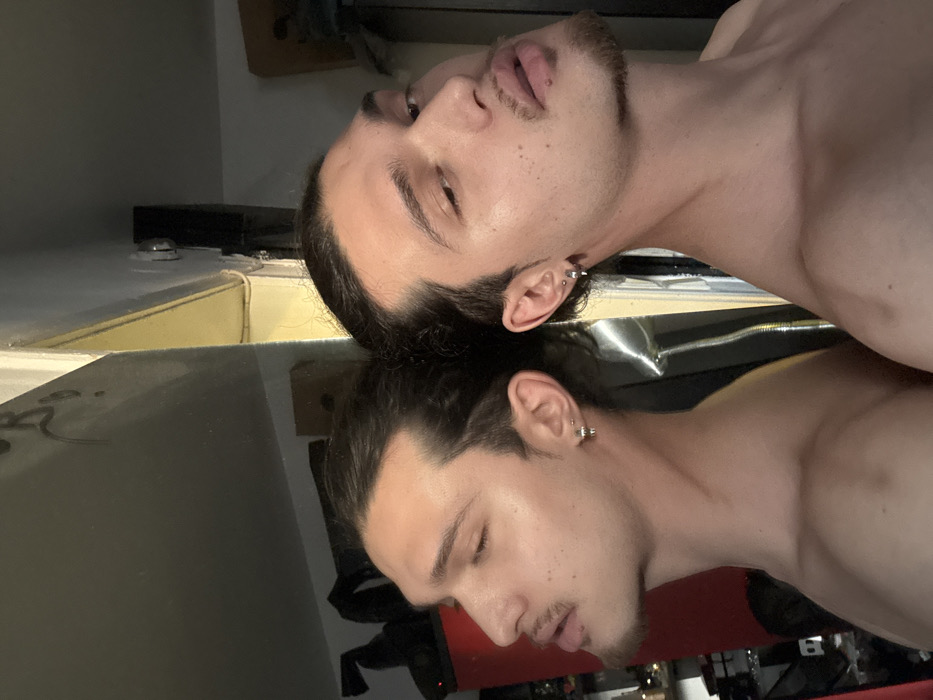 The image displays a male individual, appearing to be in his twenties or thirties, positioned shirtless in front of a large reflective surface, likely a mirror. The individual has dark hair pulled back, a mustache, and a goatee. He exhibits two silver earrings in his right earlobe and one in his left earlobe. The left portion of the image shows the individual's reflection in profile, with his eyes closed or partially closed, head tilted slightly back, and lips parted. The right portion of the image depicts the individual directly, seen from a slightly frontal angle, with eyes partially open and directed downward, head tilted slightly back, and lips parted.
The background visible behind the reflection on the left side of the image includes a red object, potentially a punching bag, and various indistinct items suggesting a cluttered indoor environment, possibly a personal room or storage area. Graffiti markings are present on the upper left section of the reflective surface. The background behind the actual individual on the right side of the image consists of a light-colored wall with a visible corner, a yellow cable or trim, and a light fixture mounted near the ceiling. The overall scene is an indoor setting. No specific identifying landmarks or features are present to determine a precise location within the city of Sofia, Bulgaria.