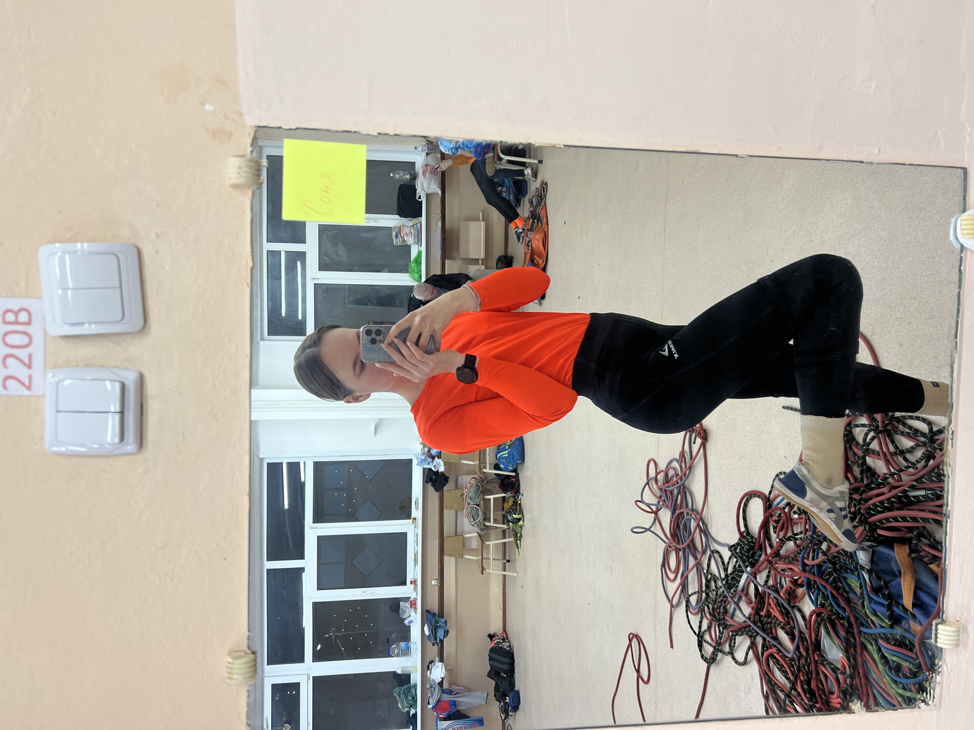 A woman is taking a selfie in a mirror in a room. She is wearing an orange long-sleeved shirt and black leggings. Her face is visible as she holds her phone up to take the picture. The mirror is reflecting a room containing numerous coiled climbing ropes, furniture, and other equipment. Through the windows in the reflection, exterior lights are visible. On the wall beside the mirror, there is a light switch and a sign reading "220B". The woman is likely standing in a climbing gym or related facility in Berezniki, Russia, based on the climbing equipment and the room layout.