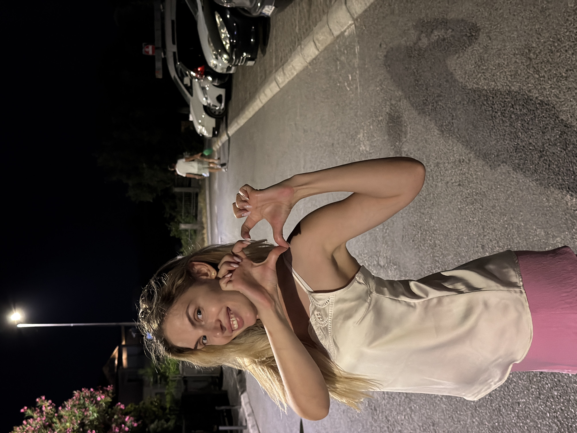 A blonde woman in a satin camisole and pink skirt forms a heart shape with her hands in front of her chest. She stands on an asphalt road at night, with cars parked along the right side of the road. A street light illuminates the scene from the left, casting a long shadow of the woman on the ground. In the background, a person walks away from the camera, carrying a green bag. Buildings and trees are visible in the distance. The location is identified as Budva, Montenegro.