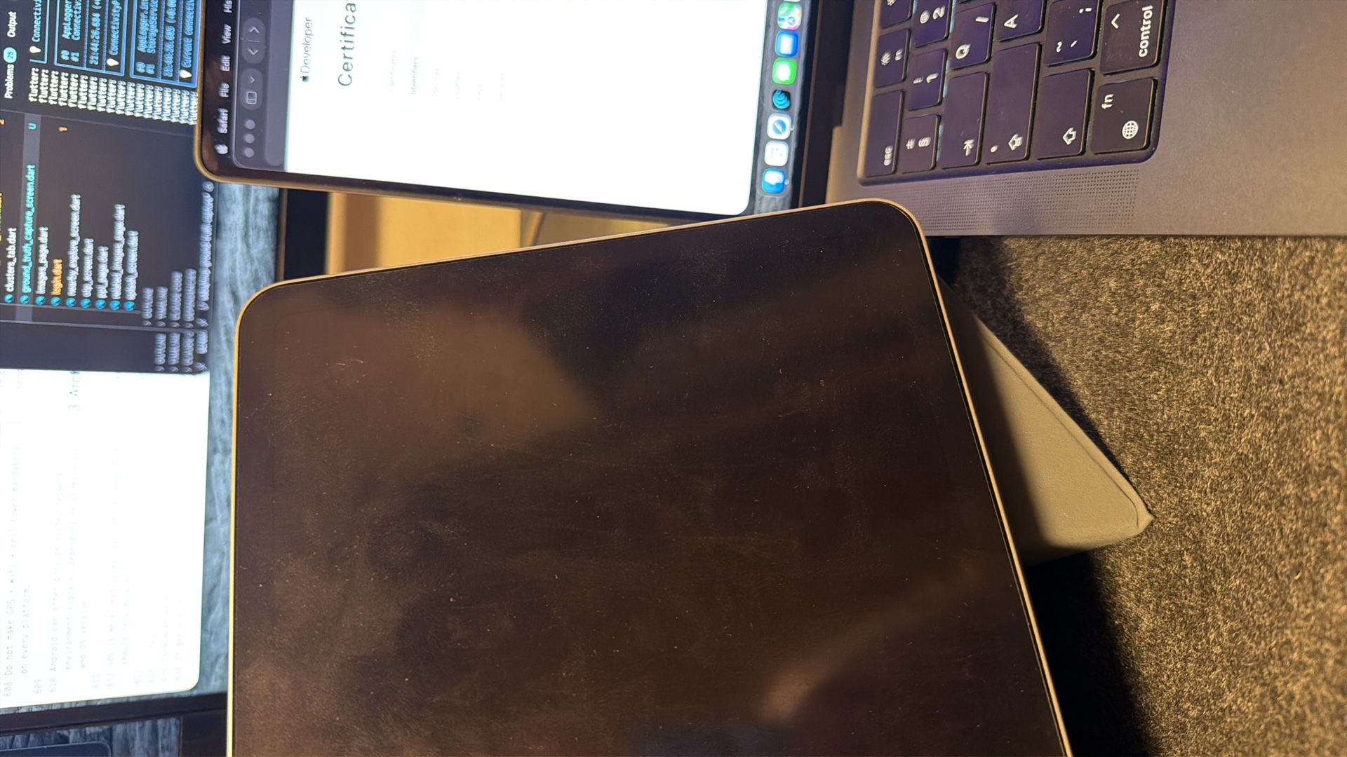 The media file is a close-up, rotated shot of a desk setup. The main subjects are a laptop and a tablet, placed on a textured, dark gray surface that appears to be a desk mat or carpet. The laptop is partially visible, showing a keyboard with dark keys and some text labels, including "fn" and "control." The laptop screen displays a white page with the word "Developer" and "Certifica" visible, suggesting a technical or coding context. To the left of the laptop, another screen is visible, displaying lines of code, with filenames like "clusters_tab.dart," "ground_truth_capture_screen.dart," and "login.dart." This screen also shows timestamps and mentions "flutter," indicating a software development environment. The tablet is positioned in the foreground, its dark, reflective surface showing some smudges and dust. A light gray, angled object, possibly a stand or a cover for the tablet, is visible beneath it. The scene appears to be indoors, likely in a workspace, with artificial lighting. There are no people or indications of time of day or weather. The visible text confirms a technical and programming-related activity.