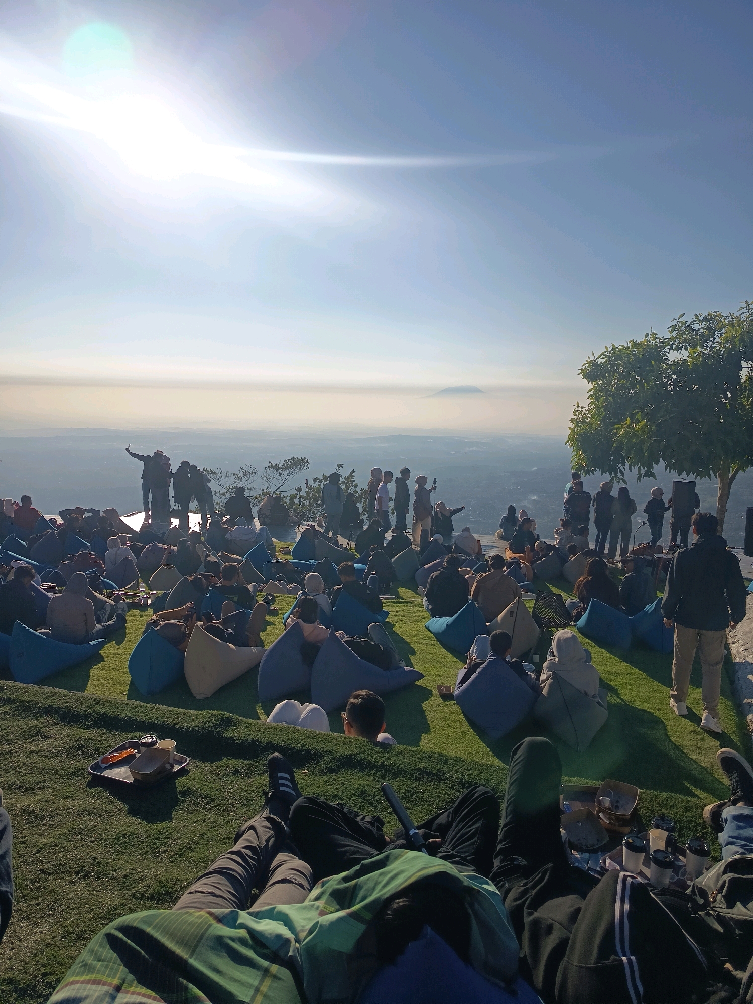 This image captures an outdoor gathering at a scenic overlook in Keditan, Indonesia, likely on a grassy knoll. A crowd of people is seated on beanbag chairs, facing a group of individuals standing on a raised platform. The standing group appears to be a band or performers, with one person holding a microphone. In the background, a vast landscape unfolds, suggesting a mountainous or hilly terrain with a hazy atmosphere, possibly indicating morning mist or distant clouds. The sun is shining brightly, creating a lens flare effect in the upper left corner of the image. Some individuals in the foreground are lounging on the grass, with personal items such as drinks and a tray scattered nearby. The overall atmosphere suggests a relaxed and enjoyable event, possibly a concert or a viewing of the sunrise or sunset.