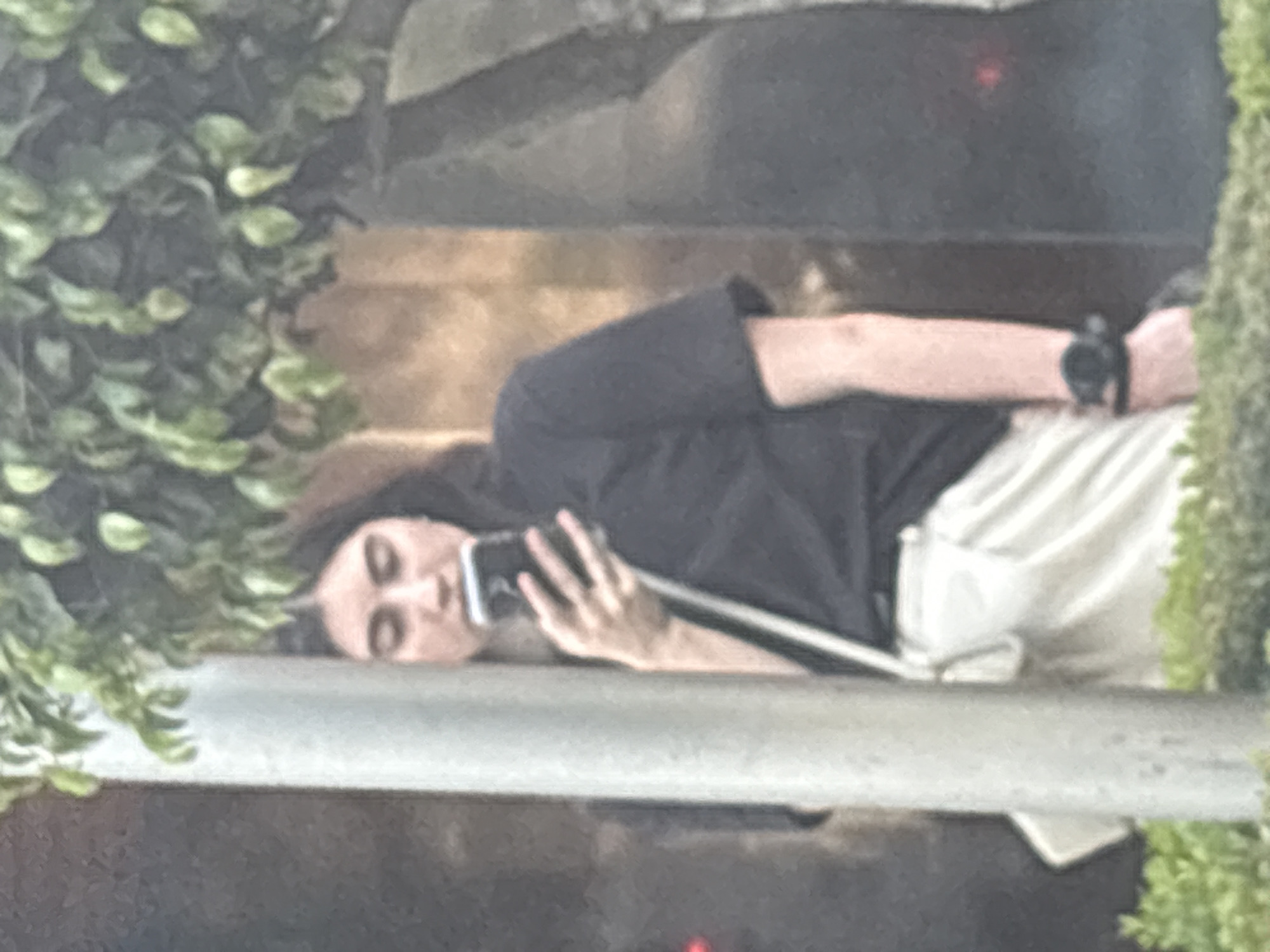 The image, taken in Mexico City, depicts a person lying down or reclining in what appears to be an indoor or shaded outdoor setting. The person, who has dark hair, is wearing a dark, short-sleeved top and light-colored trousers or pants. A black smartwatch is visible on their left wrist.
The main activity shown is the person engaging with a smartphone, which they hold with their right hand. Their gaze is directed towards the phone's screen, suggesting they might be taking a selfie, recording a video, or on a video call. Their facial expression is neutral, showing engagement with the device.
Green foliage, resembling bushes or vines, frames the scene on both sides. The background features dark, possibly wooden or concrete, surfaces, hinting at architectural elements. A light-colored railing or ledge is visible across the lower part of the frame, beneath the person. The lighting indicates it is daytime, but the immediate area is shaded. The image has a slightly soft and grainy quality. There is no visible text in the scene.