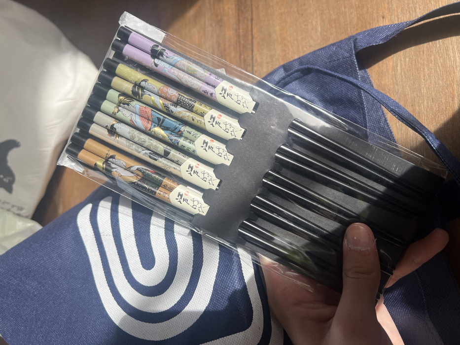 A person's left hand is centered, holding a clear plastic-wrapped package containing five pairs of decorative chopsticks. Each chopstick handle features a unique color scheme and traditional Japanese-style female figure, while the lower sections are black. The package is backed by black cardboard. To the left and beneath the package, a dark blue fabric bag with a white arched logo is partially visible. The background consists of a wooden surface and a blurry white object with an indistinct dark silhouette. Strong light illuminates the scene from the upper left. The location is the Hof Ashkelon Regional Council, Israel.