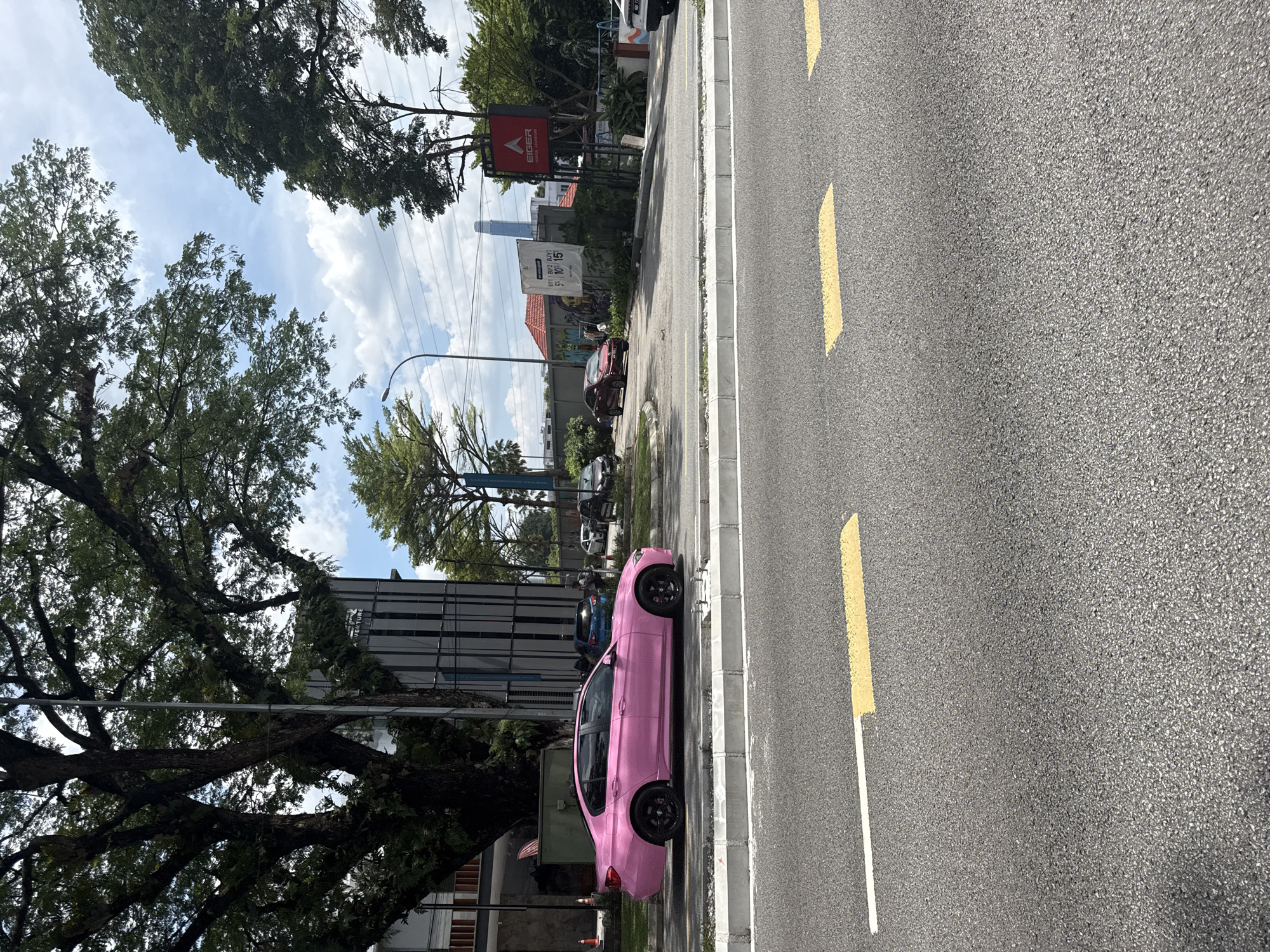 The image shows a pink BMW car parked on the side of a road in Kuala Lumpur, Malaysia. The road has yellow dashed lines marking lanes. On the side of the road, there's a sidewalk and various trees. In the background, there are buildings, including the Exchange 106 skyscraper. A sign for Eiger Adventure can also be seen in the background.