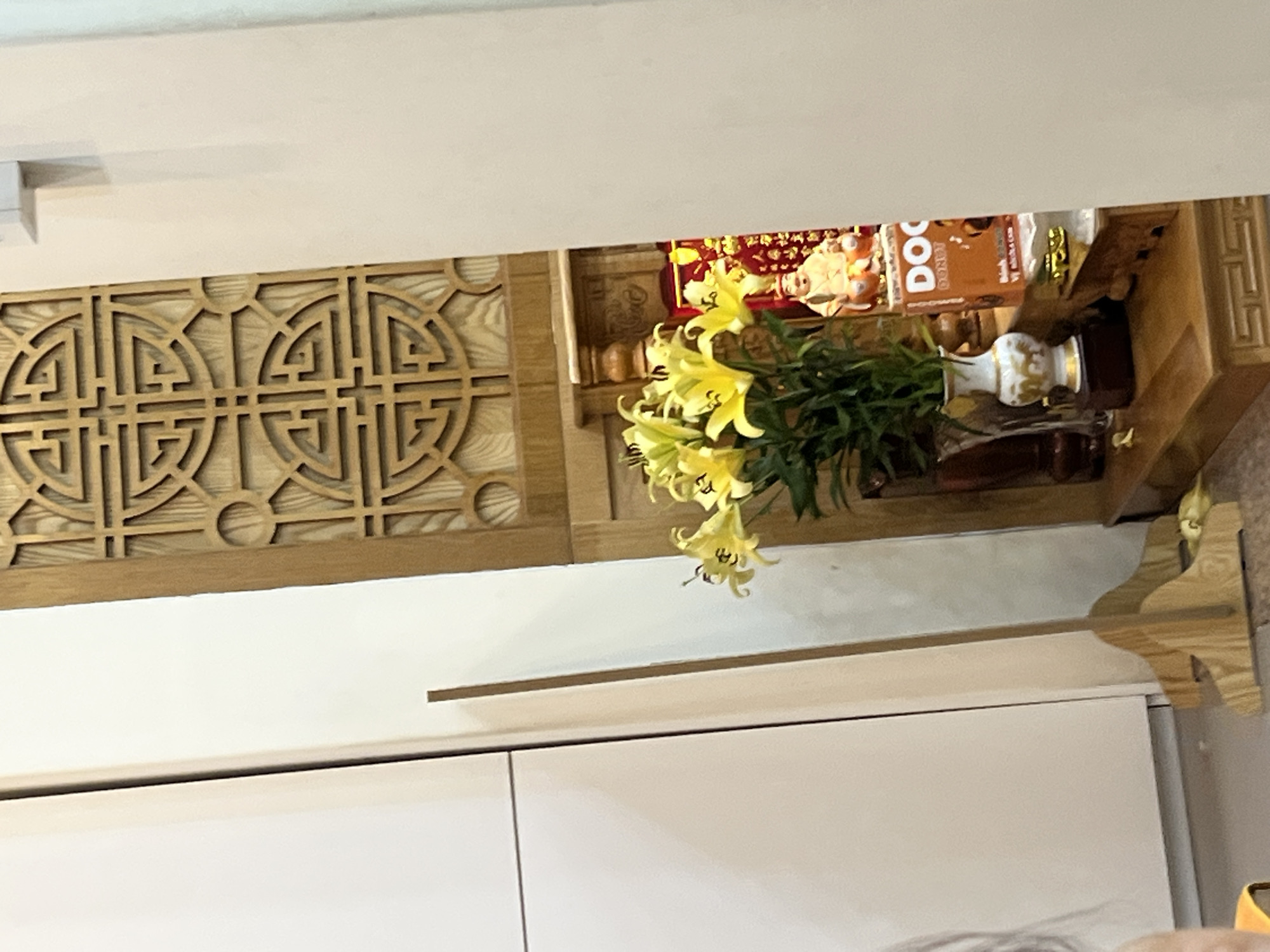 An indoor scene shows a religious altar with a wooden screen, yellow lilies in a vase, and a small statue. To the left is a white cabinet. The altar is made of wood and decorated with a traditional motif. The yellow lilies are in a white and gold vase placed on a wooden stand. Behind the vase is a small statue of a figure, possibly a deity, and an orange box with the word "DO" visible. The overall impression is of a domestic religious setting. The exact location in Nha Trang Ward, Vietnam, is not specified within the image itself, but it is an interior view.