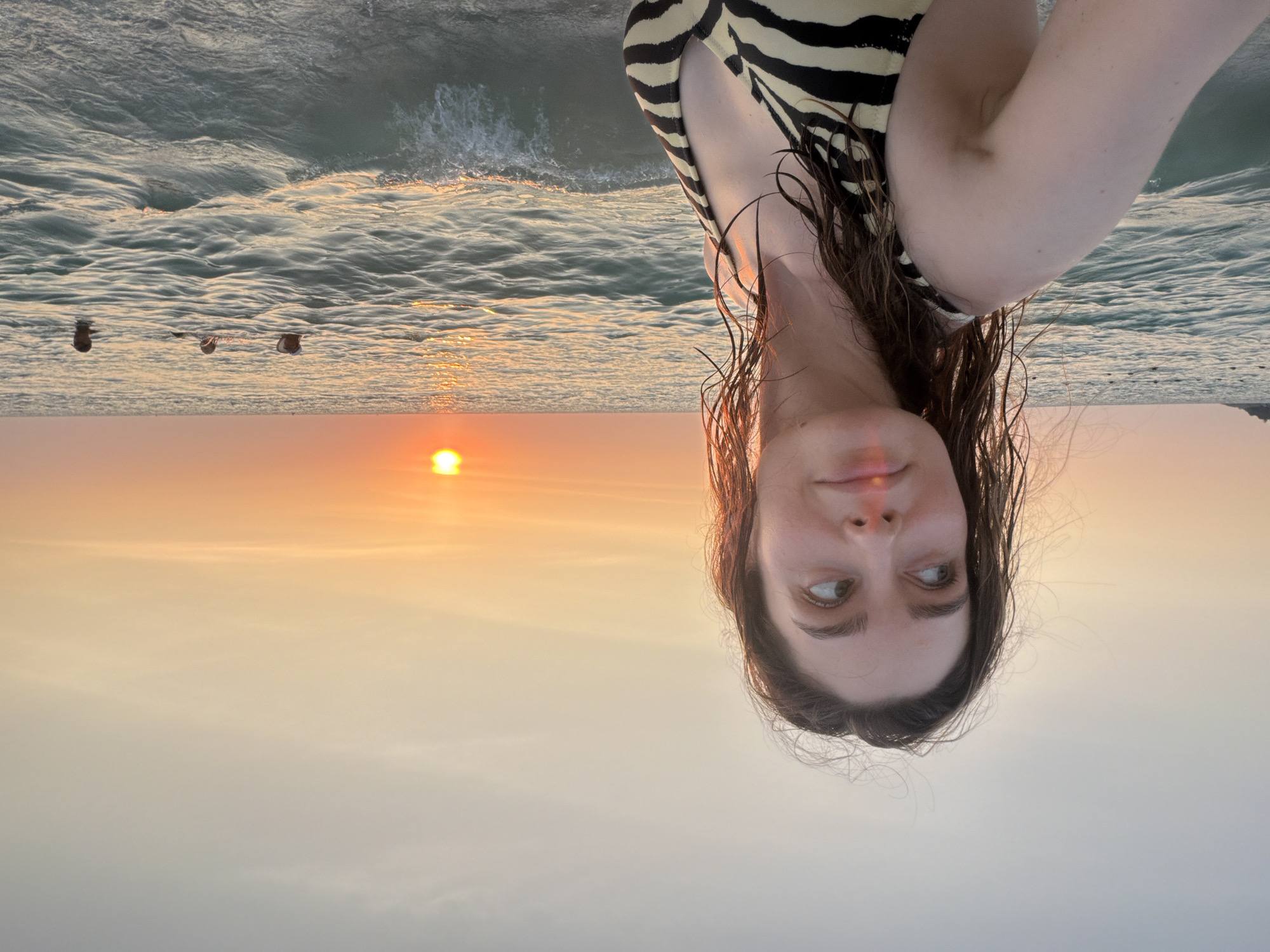 The image depicts a sunset scene at Karon Beach, Karon, Thailand. In the foreground, a fair-skinned young adult female with wet, dark brown hair stands in shallow ocean water, wearing a black and light-yellow zebra-print bikini top. She is turned slightly towards the viewer but gazes to her right (viewer's left), with a faint smile. The setting sun, a prominent orange disc, is visible low on the horizon in the upper right quadrant, casting an orange-yellow glow across the sky and the rippling ocean surface. The sky transitions from a vibrant orange at the horizon to a lighter, hazy blue-grey overhead. Small waves are breaking in the foreground. In the mid-ground, three additional individuals are partially submerged in the ocean water. To the far left, a dark landmass, possibly a distant headland or island, is visible on the horizon, accompanied by several dark, spherical objects floating in the water, likely buoys or markers.