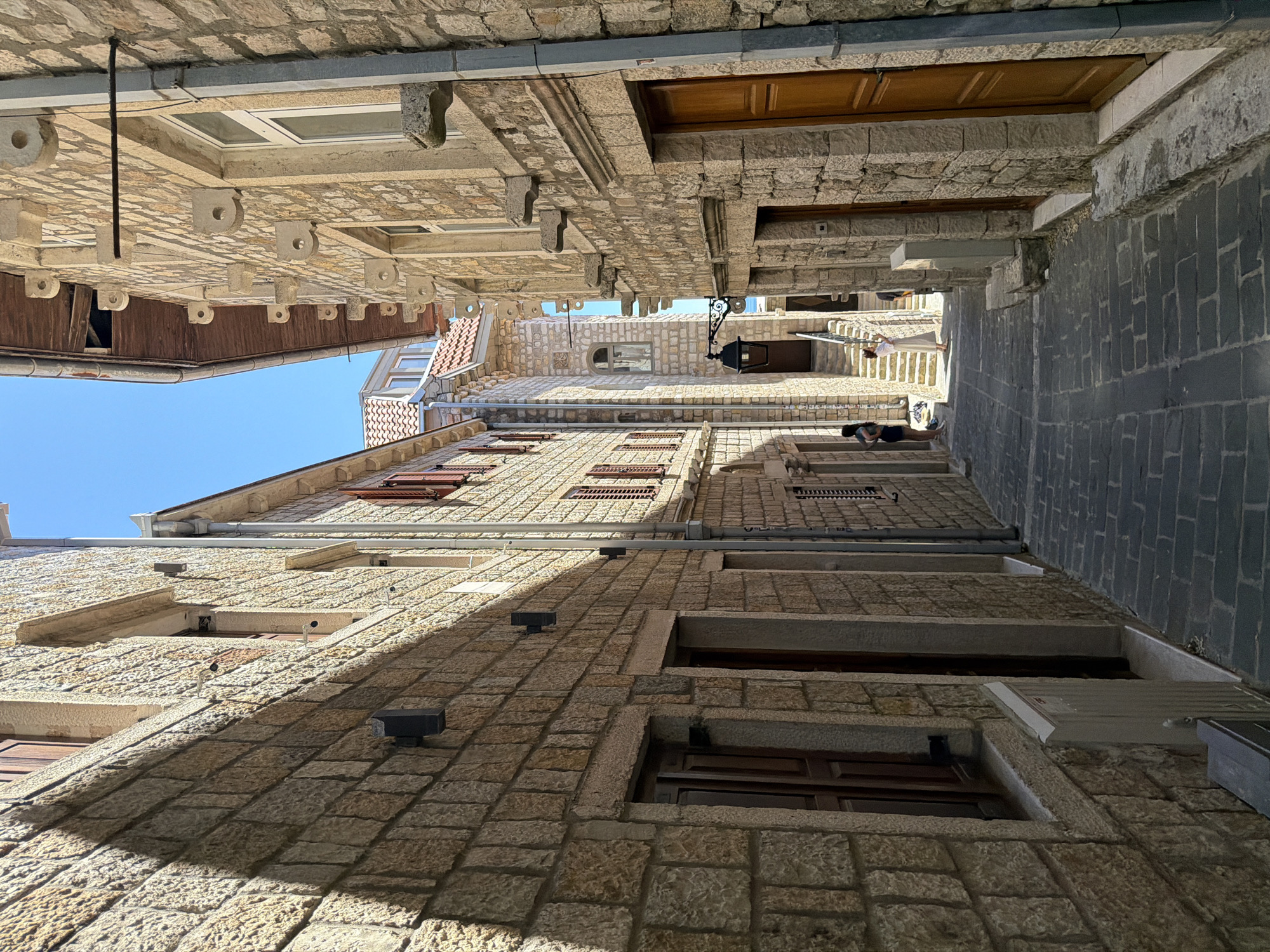 This is a street-level, eye-level shot of a narrow cobblestone alleyway in the old town of Ulcinj, Montenegro. The architecture is characterized by weathered stone buildings with wooden doors and shutters, reflecting a historical Mediterranean style. Sunlight casts strong shadows, emphasizing the texture of the stone walls and the unevenness of the paving.
On the left, a stone building with visible brickwork features two darkened wooden doors, one partially obscured by a metallic gray utility box. The facade is adorned with two black exterior lights. Above, a window with wooden shutters is visible, casting a shadow on the wall.
The right side of the alleyway is dominated by a stone building with a warm, brown wooden door. Above the door, a row of peculiar, circular stone decorative elements are mounted along the building's facade. A metal pipe runs vertically down the side of this building.
Further down the alley, the path slopes upwards towards a set of stairs. At the base of the stairs, two women are visible. One, on the left, is wearing a dark top and shorts, standing in a shaded area. The other woman, on the right, is wearing a white dress and appears to be interacting with the stairs or the area ahead. Another person is partially visible on the stairs. A vintage-style lantern hangs from a wrought-iron bracket on the wall, adding to the old-world charm.
The sky overhead is a clear, bright blue, indicating a sunny day. The overall impression is one of an ancient, sun-drenched European town, likely a popular tourist destination given the presence of pedestrians.