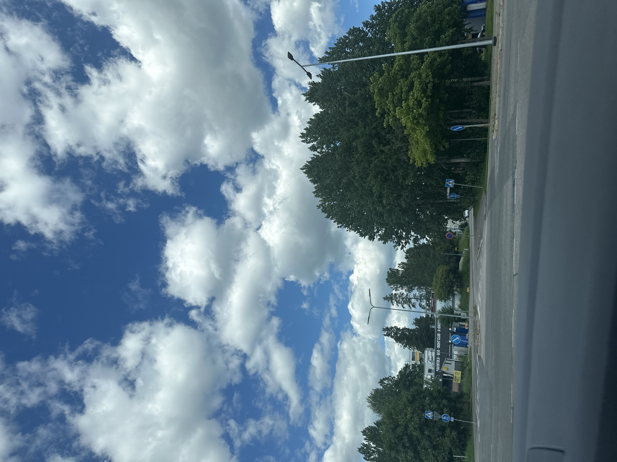 The image shows a street view of an intersection in a city, with a bright blue sky filled with fluffy white clouds. Tall trees with dense green foliage line the right side of the road, and a street light is visible. On the left side of the intersection, there are buildings, including one with text that reads "PAVINTOLA - BAR - NIGHT CLUB - A-OIKEU". Various traffic signs are present, including pedestrian crossing signs and directional signs. The road surface is asphalt with white lane markings. The bottom of the image shows the dashboard of a vehicle, indicating the photo was taken from inside a car.