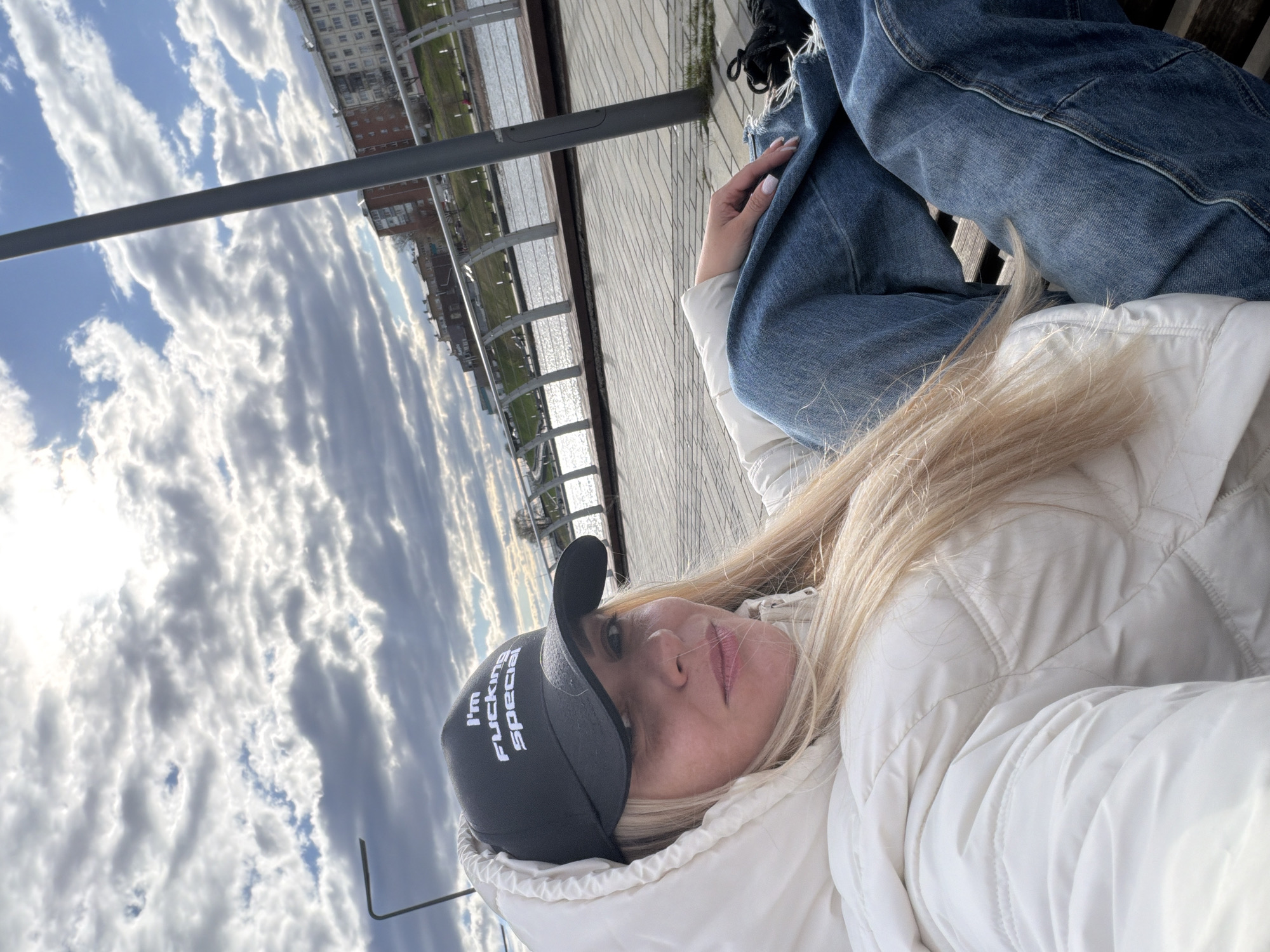 The image shows a woman wearing a white puffy jacket and a black baseball cap with the words "I'm Fucking Special" printed on it. She is lying down, seemingly on a bench, with her head tilted back. Her blonde hair is spread around her face and shoulder. She is also wearing blue jeans.
In the background, there is a river or body of water. Beyond the water, there are buildings visible. The sky is cloudy with patches of blue visible between the clouds. A metal railing is present along the edge of the walkway or bench area.
Based on the buildings and the river, it is possible this image was taken in Votkinsk, Russia, but it's not definitively possible to confirm with certainty.