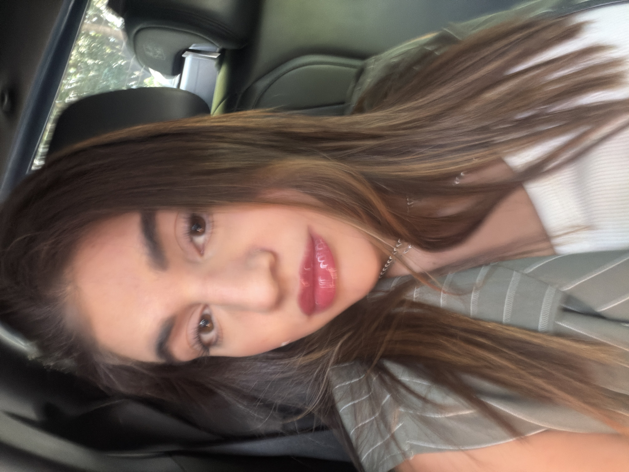 A medium shot of a woman with long brown hair and fair skin, wearing a light green and white striped shirt. She is in a car, with the headrest of a black leather seat visible behind her head. The car window shows trees in the background. The location is unknown.