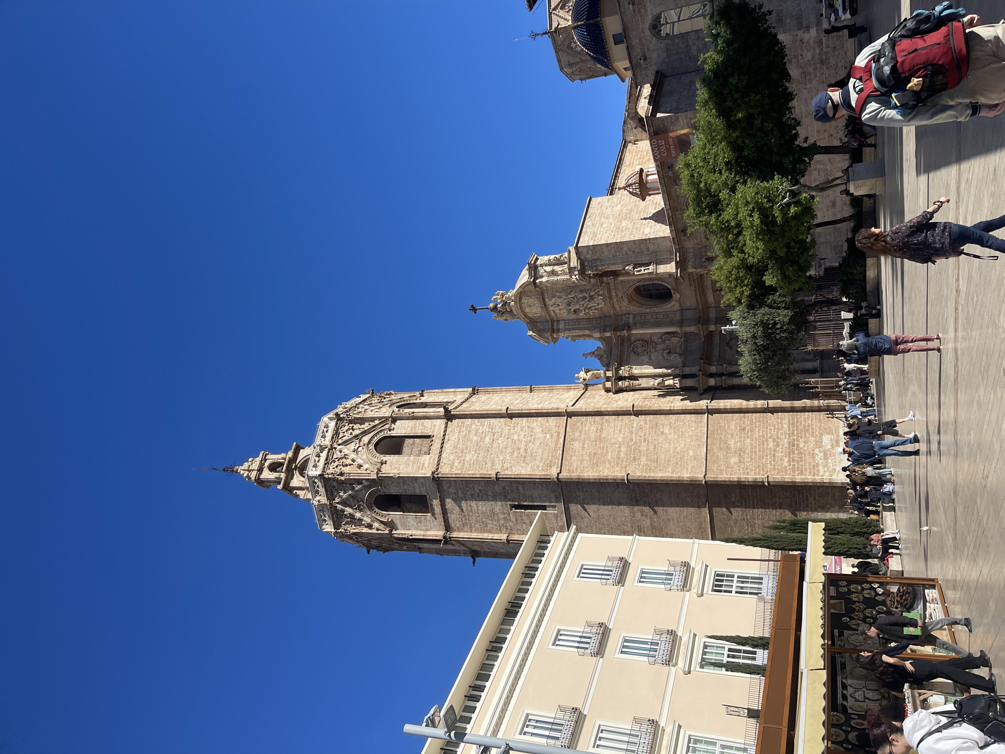 The image depicts the Micalet bell tower and Valencia Cathedral complex in Valencia, Spain, under a clear blue sky. The stone bell tower rises prominently, connected to adjacent buildings. Below, a plaza is populated with pedestrians walking and standing near market stalls. A light-colored building with multiple windows and ornate iron balconies anchors the lower portion of the frame.