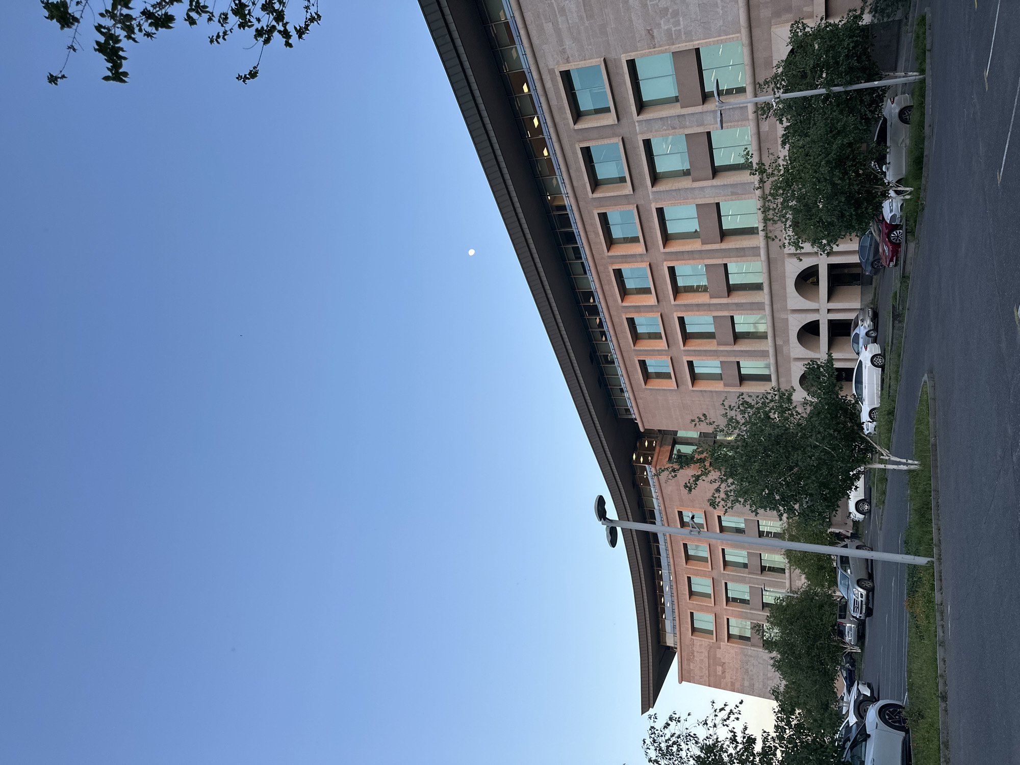 The image shows a large, rectangular building with a repeating grid of windows under a clear blue sky. The building appears to be made of stone or a similar light-colored material. There are trees in front of the building, obscuring some of the lower levels. Cars are parked along the side of the building, on a paved road. A streetlight is visible near the building. In the sky, a small, pale circle resembling the moon is present. While no people are visible, the presence of parked cars indicates human activity. The location is identifiable as Yerevan, Armenia based on the architectural style of the building, which is similar to structures in the city center.