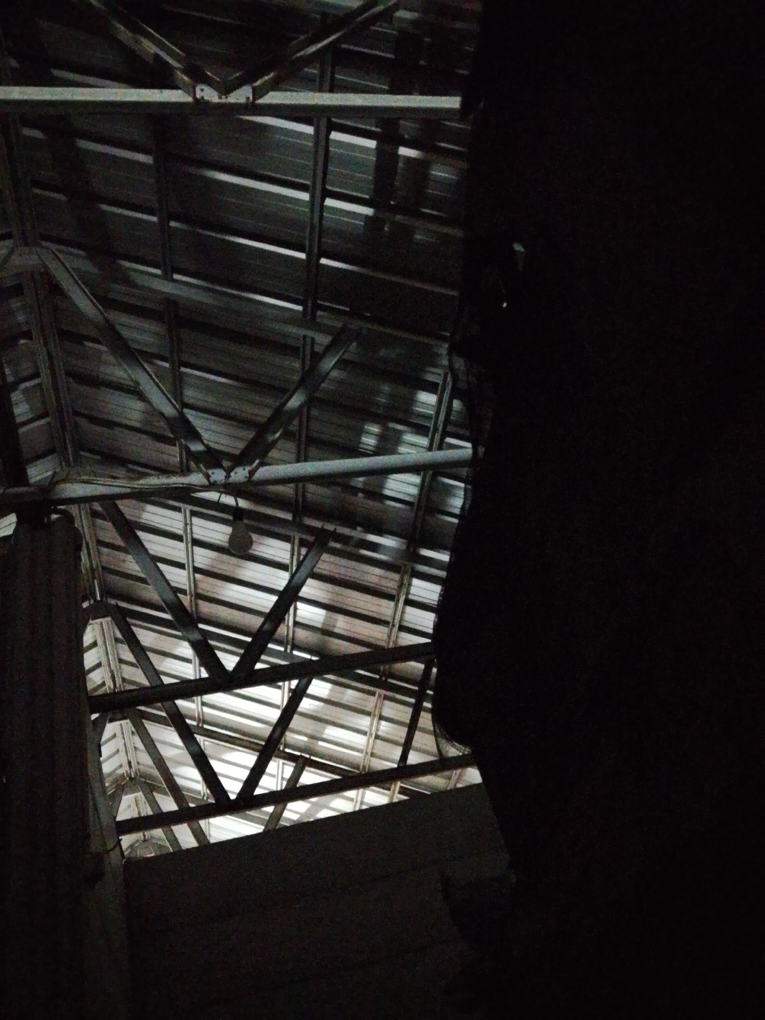The image, taken in Bukit Ketok, Indonesia, shows the interior of a structure with a metallic corrugated roof supported by steel beams. A bare lightbulb hangs from the structure. The right portion of the image is dominated by a large, dark, out-of-focus shape. A section of wall is visible in the lower left. The overall lighting is dim.