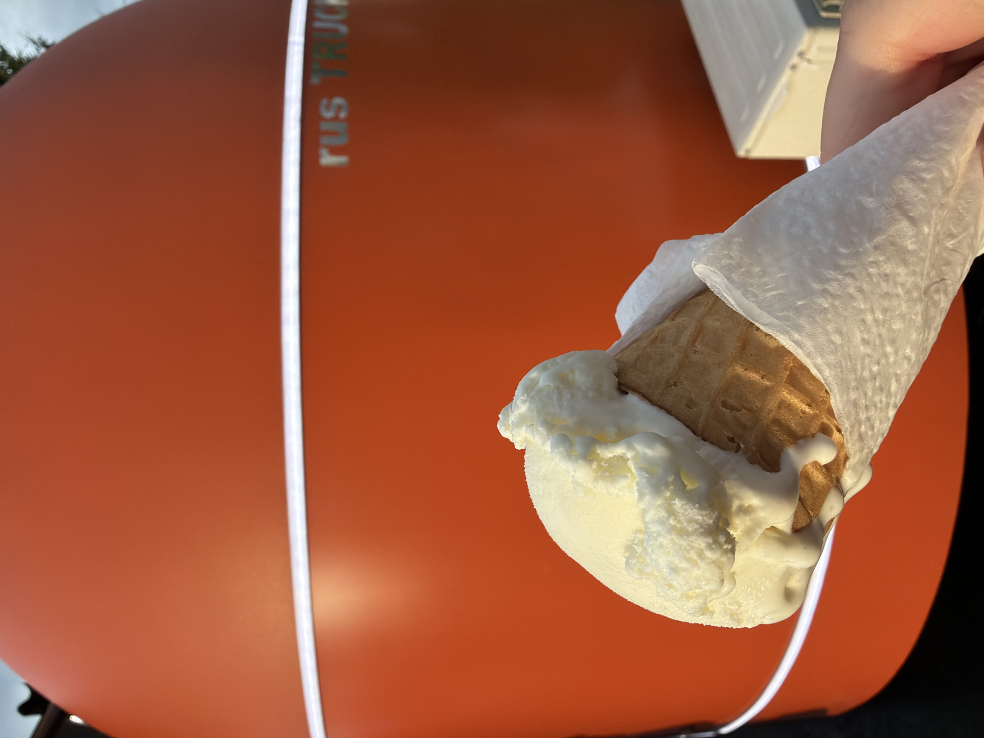 A person's hand holds a vanilla ice cream cone in front of an orange, rounded structure. The ice cream is melting, with drips of white cream running down the side. The cone appears to be a standard waffle cone, wrapped in white paper. The orange structure has a white band of light circling it and the words "rus TRUCK" are visible in silver lettering on its surface. In the background, a white rectangular object, possibly part of the truck or another piece of equipment, is visible. The scene appears to be outdoors, with hints of a cloudy sky and foliage in the upper left corner. The location is Unknown, Unknown.