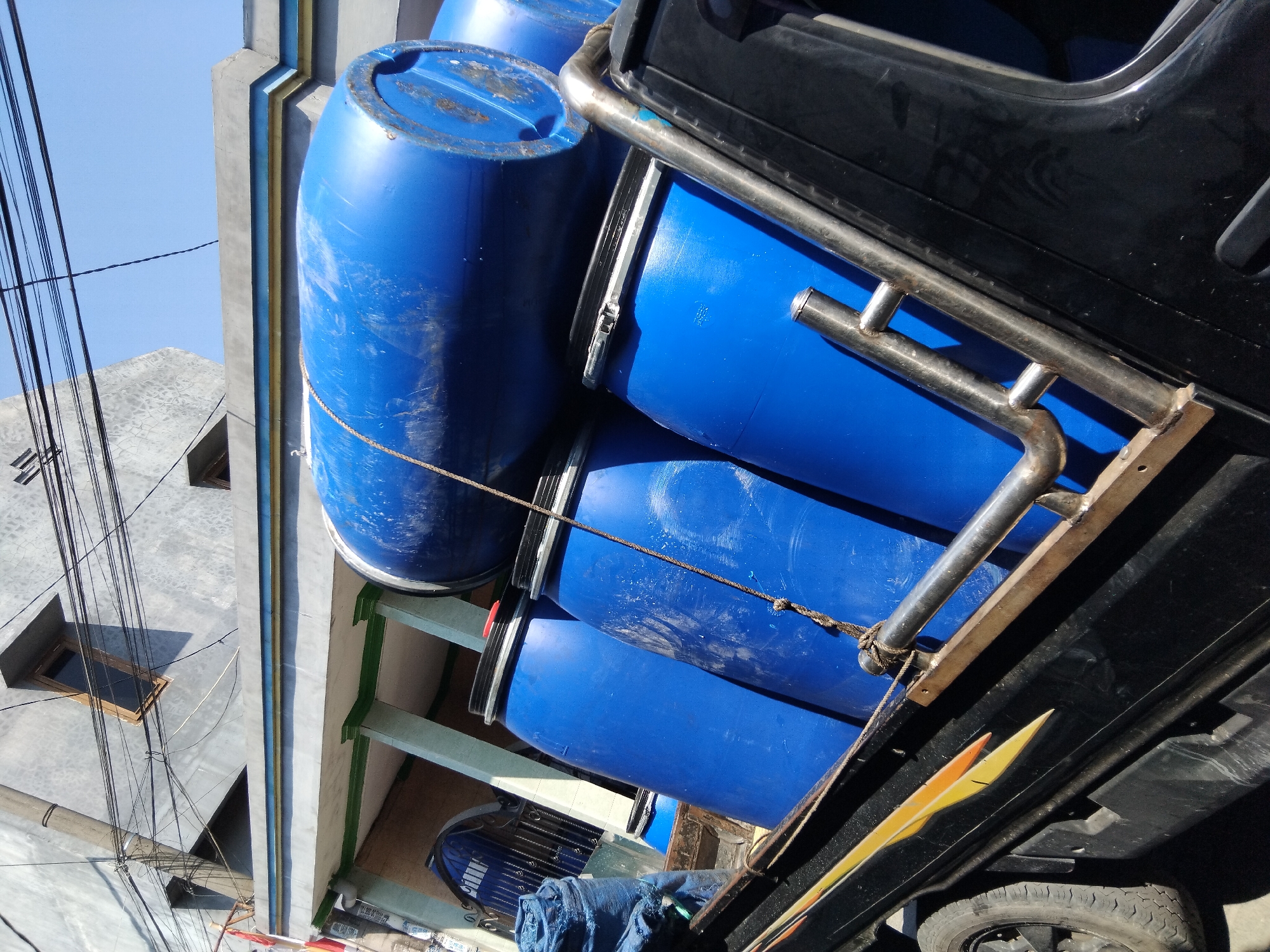 A flatbed truck is loaded with multiple large blue plastic barrels, secured by a rope. The truck bed features decorative yellow and orange flame-like decals. In the background, a multi-story building with grey walls and windows is visible, along with an overhead network of electrical wires. The scene appears to be located in Kepuh Kiriman, Indonesia, indicated by the presence of an Indonesian flag on a pole attached to the building. The sky is clear and blue.