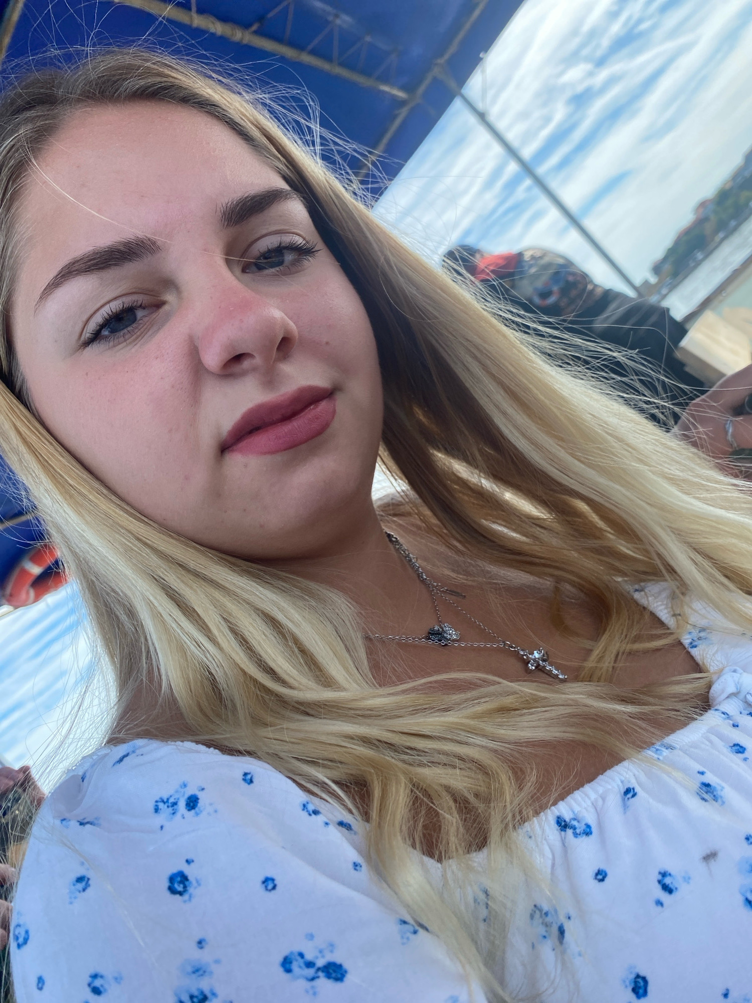 The image is a close-up, low-angle selfie of a young woman with blonde hair and light skin, taken outdoors. She is wearing a white top with a blue floral pattern and a silver necklace with a cross pendant and a small heart charm. Her makeup includes eyeliner and pink lipstick. Behind her, the sky is blue with wispy white clouds. A blue awning or canopy is visible above, and parts of a boat or vehicle are present. The background also shows a body of water and a distant shoreline with buildings, suggesting a coastal or waterfront location. The lighting is natural and bright, indicating it is daytime. The image appears to be taken on a boat or near the water, possibly in the city of Nesebar, Bulgaria, given the architectural style of the distant buildings and the general coastal setting.