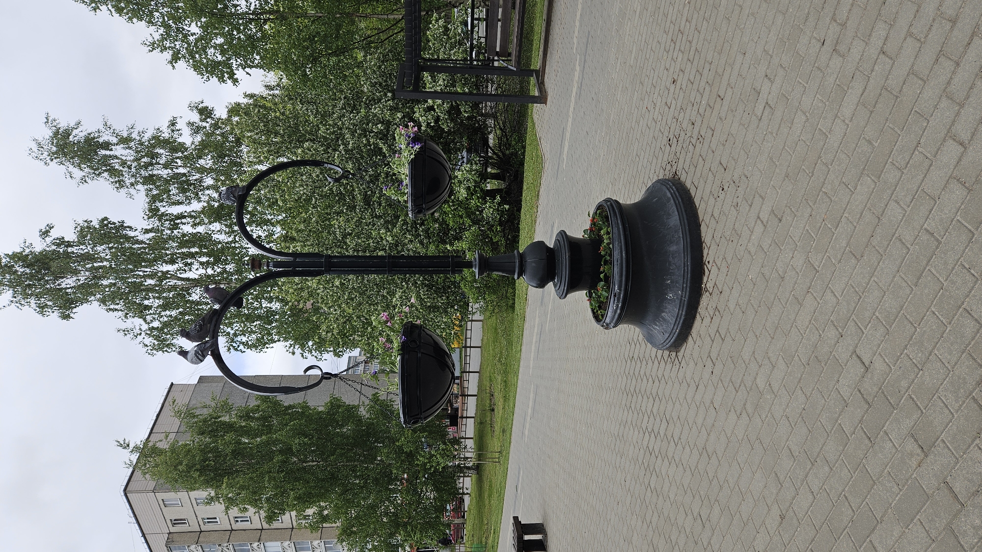 The image shows a black, ornate streetlamp in a public park. The base of the lamp is a large, decorative pot filled with red flowers. Two curved arms extend from the lamp, each supporting a hanging flower basket with purple flowers. Several pigeons are perched on the lamp's arms. The background features a paved walkway, green grass, trees, a bench, and a multi-story building. The sky is overcast. The location appears to be a city park in Unknown, Unknown.