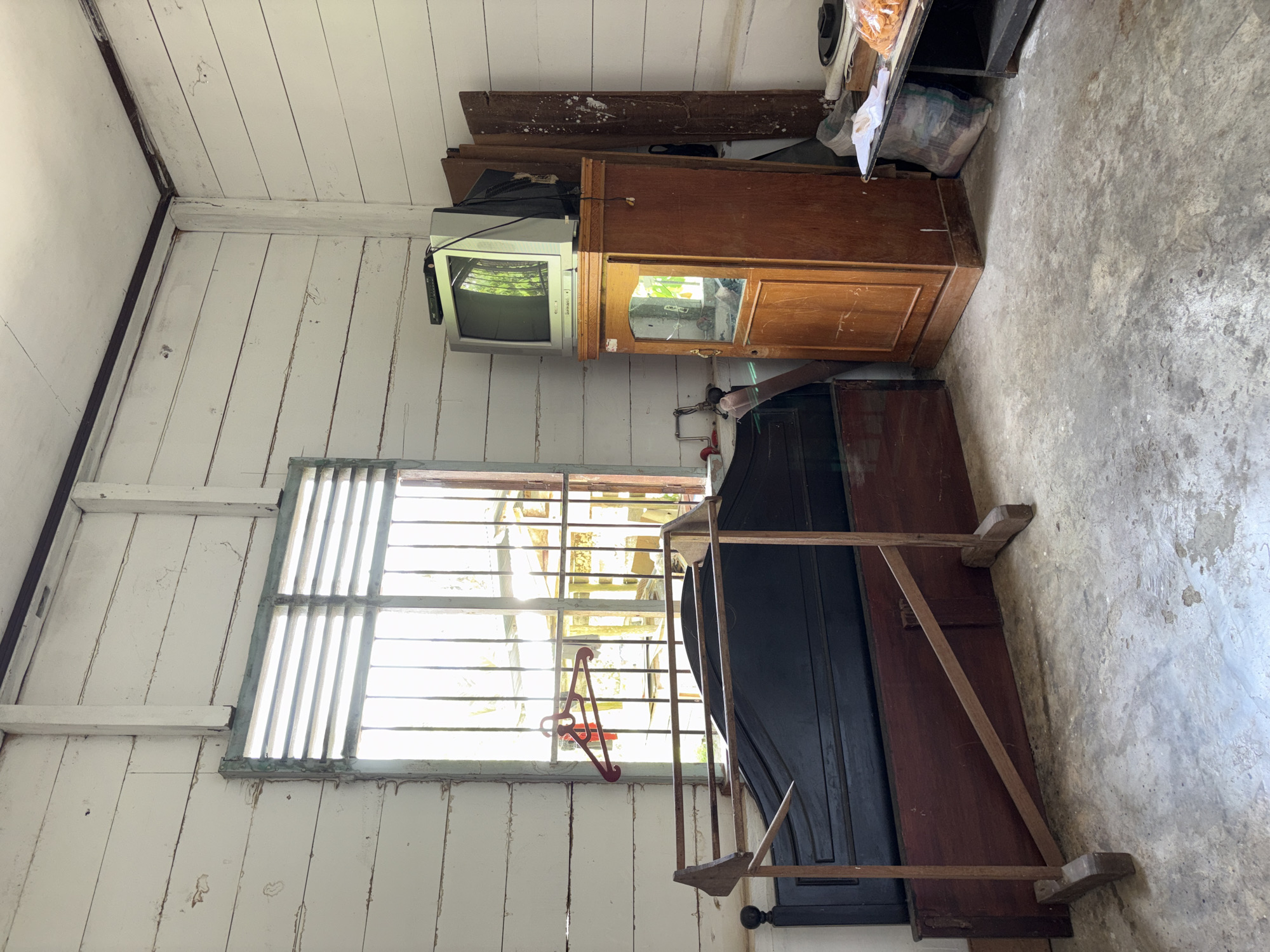 The image depicts the interior of a room with walls and ceiling made of white-painted horizontal wooden planks. A large window with horizontal louvers and bars is visible on the left, allowing sunlight to enter and illuminating the room. In front of the window, a black headboard of a bed is present, with a dark reddish-brown wooden base. To the left of the bed, a wooden clothes rack with an angled support is set up. A red clothes hanger hangs from the window bars.
On the right side of the room, a tall, narrow wooden cabinet with a glass door stands. A vintage television set, with a visible VCR or similar device on top, is placed on the cabinet. Behind the cabinet, there are several wooden planks leaning against the wall, and to the right of the planks, a small black table holds a plastic bag, some papers, and a bag of snacks. The floor is made of concrete and shows signs of wear and staining. The overall impression is that of a simple, somewhat aged room. The location is identified as the city of Beunyot, Indonesia.