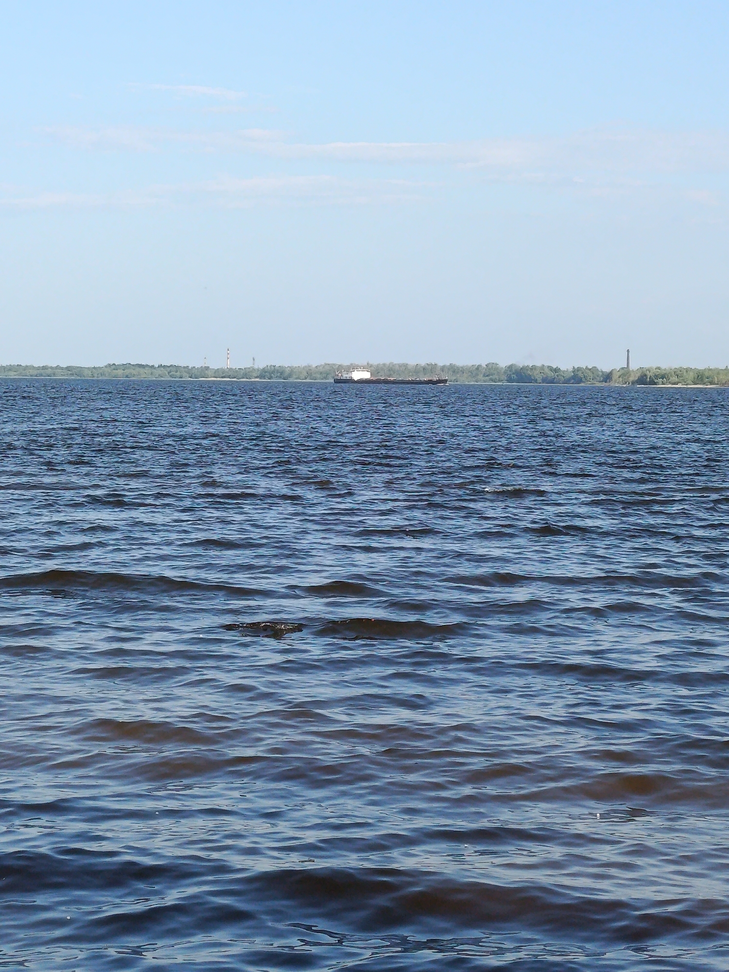 A wide shot shows a calm river under a clear blue sky with a few wispy clouds. In the distance, a dense line of green trees forms the opposite bank. Two tall, slender industrial chimneys are visible on the distant shore, one with red and white markings. A large, white barge with a dark hull is moving across the water in the middle distance. The foreground is dominated by the rippling surface of the river, reflecting the light and creating a textured pattern of blues and grays.