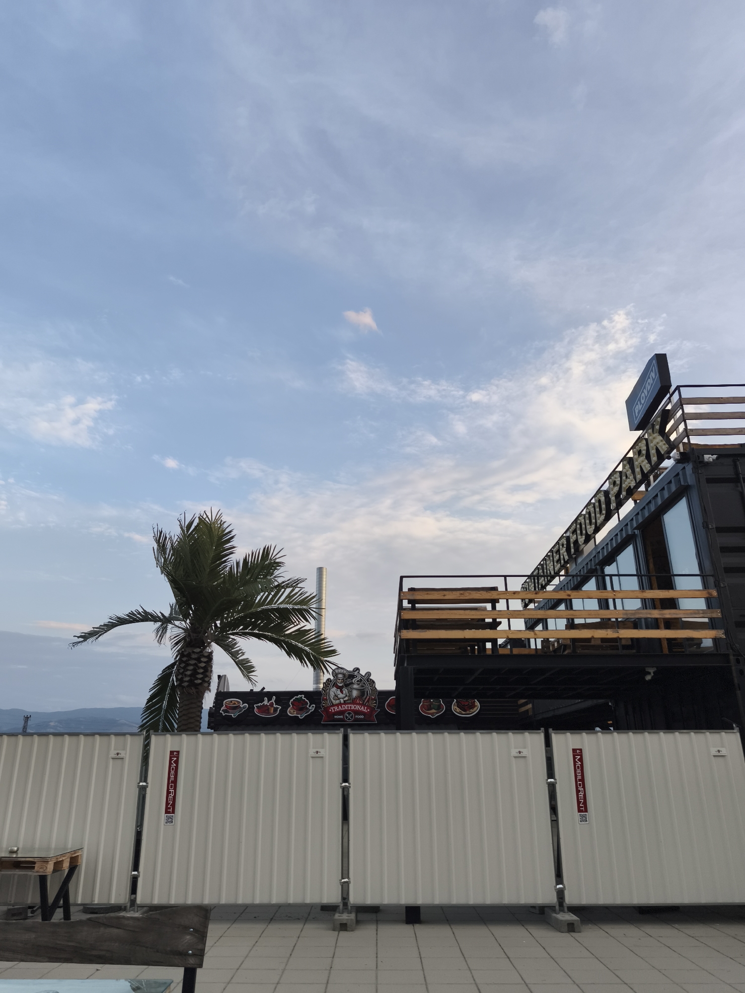 The image depicts an outdoor scene in Plovdiv, Bulgaria, featuring a modern structure built from shipping containers, likely a food park, with a sign that reads "PLOVDIY FOOD PARK". The structure has multiple levels, including a balcony area with wooden railings. A single palm tree stands to the left of the container building, its fronds reaching towards the cloudy sky. In the foreground, a white corrugated metal fence separates the viewer from the food park. The fence has several metal posts and appears to be temporary, possibly for construction or an event. There are also some wooden elements and a bench visible in the lower left corner, suggesting an outdoor seating area. The sky is partly cloudy with a soft, diffused light, indicating it could be early morning or late afternoon. In the background, distant hills are visible beneath the horizon line. The overall impression is of a contemporary urban space designed for dining and socializing.