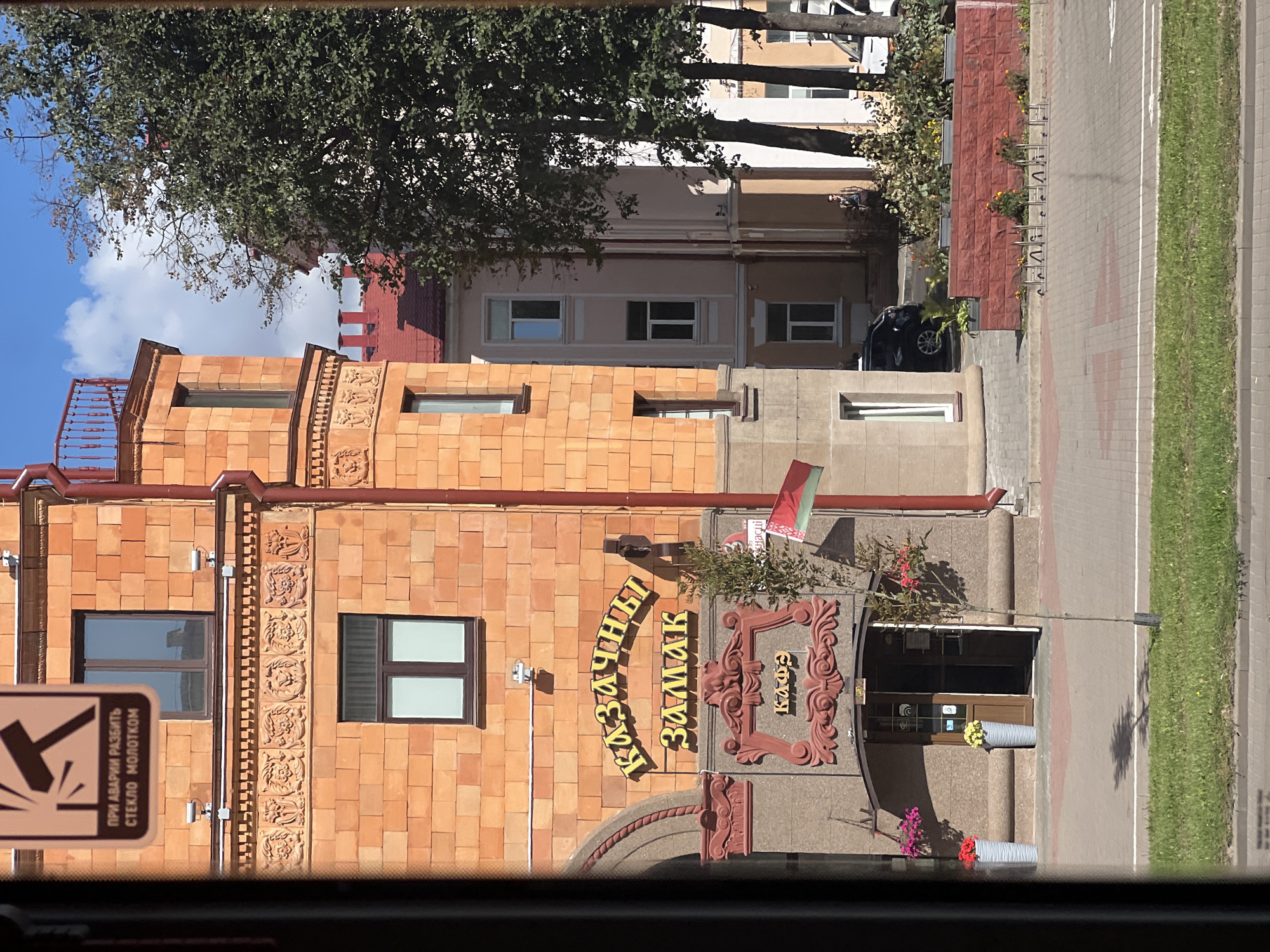 The image captures a view through a window onto a street in Minsk, Belarus. The main focus is an orange brick building with a prominent arched sign reading "Казачья Замакъ" in yellow stylized Cyrillic letters. Below this, a decorative red relief surrounds another sign that says "Кафе". The entrance to the cafe is visible, with an open door and an awning. Two large grey planters with colorful flowers flank the entrance, and a young tree stands to the right. A flag, likely the flag of Belarus with its red and green stripes, hangs from a pole on the building.
Behind the orange building, a lighter-colored building with multiple windows is visible. Trees with green foliage line the street, and a red brick planter wall is present in the mid-ground. A black car is parked on the street. The overall scene suggests a pleasant, sunny day in a city.