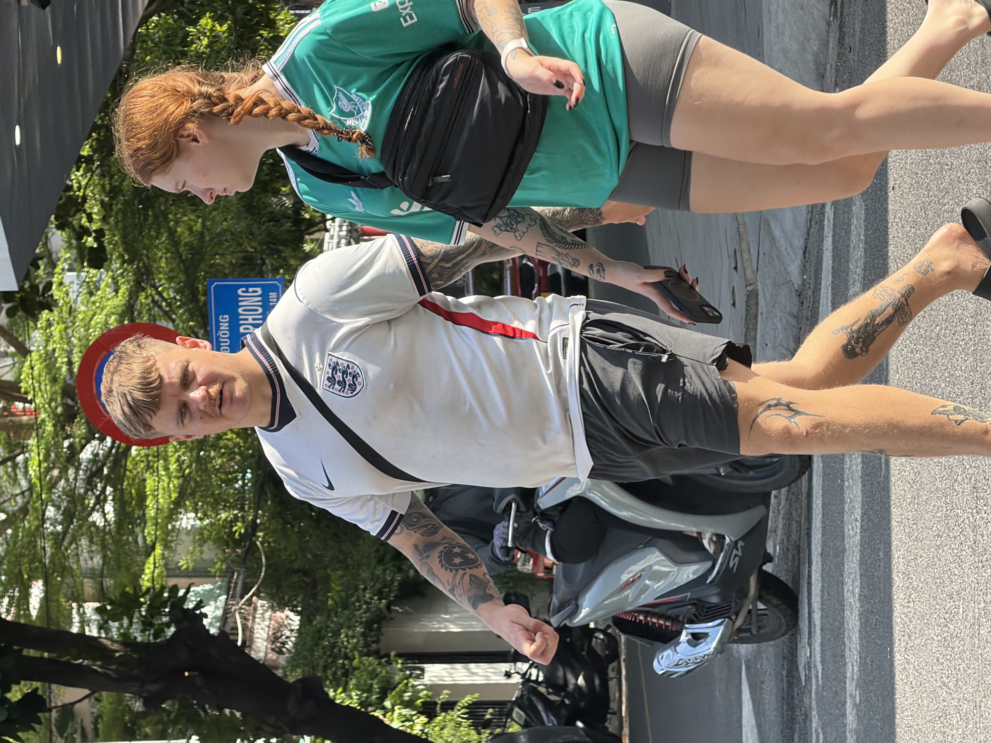Two individuals, a male and a female, are walking on a street in Nha Trang Ward, Vietnam. The male, on the left, is wearing a white England soccer jersey and dark shorts. He has numerous tattoos on his arms and legs. The female, on the right, is wearing a green soccer jersey and grey shorts, with her hair in a braid. She has a black fanny pack across her chest and tattoos on her arms. They are walking on an asphalt road, with a motorcycle parked to the left of the male. In the background, there are trees, buildings, and a street sign with Vietnamese text. The scene appears to be an outdoor, daytime setting.