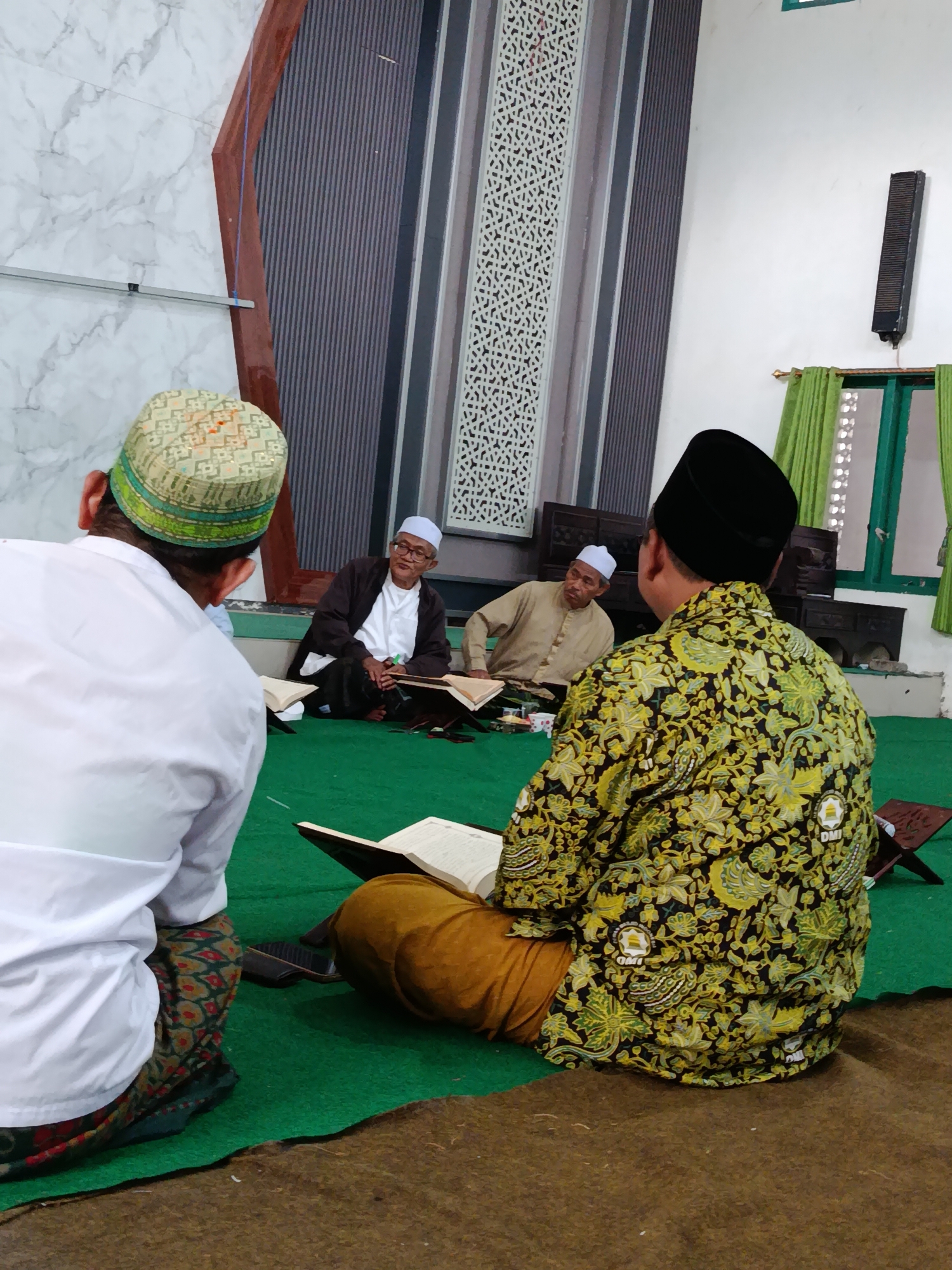 In the city of Bondowoso, Indonesia, four individuals are seated on a green carpeted floor in what appears to be a prayer room or religious gathering space. The room has marble-patterned walls on the left and vertically striped wall panels on the right. In the background, there are wooden benches and a window with green curtains. The men are all wearing traditional Islamic attire.
In the foreground, facing away from the camera, is a man in a white long-sleeved shirt and a patterned sarong. He wears a green and white patterned skullcap. He is looking towards the other three individuals who are positioned further into the room.
To the left of the man in white, and slightly behind him, is another man wearing a white skullcap and glasses, a brown outer garment, and dark pants. He is looking towards the center and appears to be engaged in reading from an open book placed on a wooden stand in front of him.
In the center background, another man wears a white skullcap and a light brown long-sleeved shirt. He is also looking towards the center, and an open book is visible in front of him.
On the right side of the image, a man is seen from behind, wearing a black skullcap and a vibrant yellow and black patterned shirt. He is also seated on the green carpet, with an open book on a wooden stand in front of him. His sarong is a mustard yellow color.
The overall scene suggests a religious lecture, study session, or prayer gathering, with the men engrossed in their books and the spiritual activity.