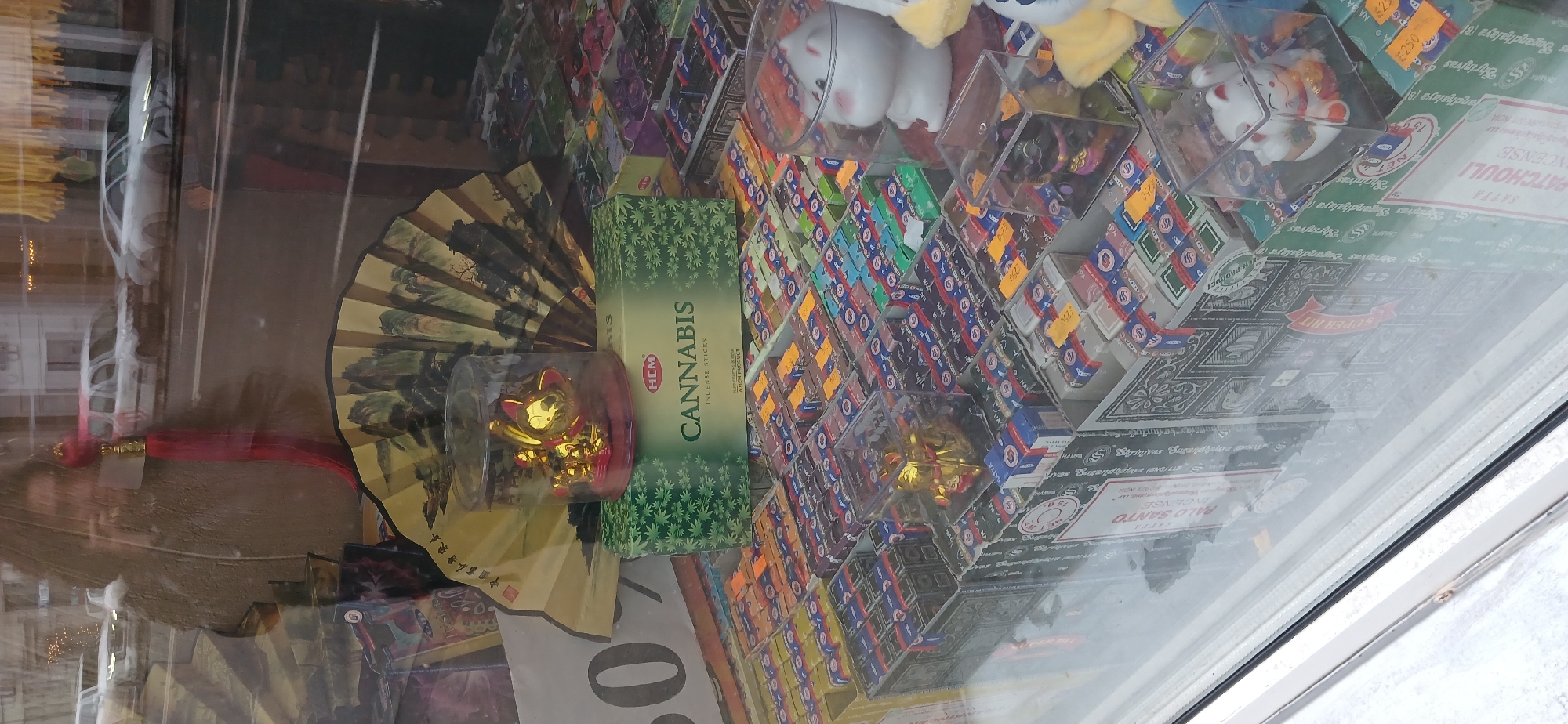 This image shows a storefront display inside a business, likely in Moscow, Russia. The display includes a variety of items for sale, including: a decorative folding fan with traditional Asian imagery, a plastic container holding a gold-colored cat figurine (possibly a "lucky cat"), boxes of "Cannabis" scented incense sticks, numerous small boxes of other incense varieties (with price tags indicating a cost of "£250"), and several plush toys of cats and other creatures in clear plastic display boxes. The incense is from brands such as "HEM", "Shrinivas Sugandhalaya", and "SATTA". The text "PALO SANTO INCENSE" and other text in English are also visible on the incense packaging. The store appears to be reflected in the glass of the display window. The image is rotated clockwise, and the orientation should be corrected.