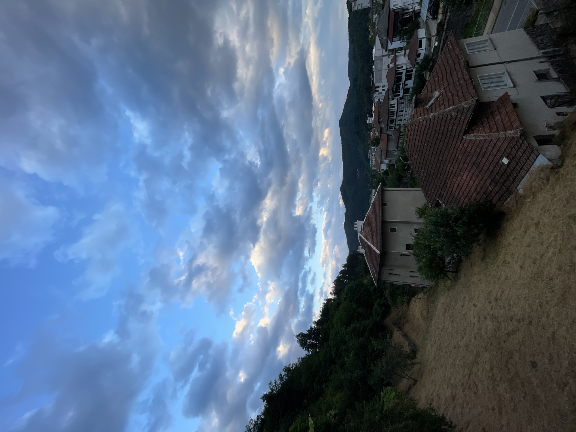 The image captures a view of Zlatograd, Bulgaria, on an overcast day with dramatic clouds filling the sky. In the foreground, a steep, dry, grassy slope descends from the left. Two houses are visible on the slope. The upper house is a light-colored, two-story building with a reddish-brown tiled roof and several windows. The lower house is partially obscured, but appears to be an older structure with a similarly colored tiled roof and stone foundation.
Beyond the houses, the town of Zlatograd unfolds. Numerous buildings with red and white facades and tiled roofs are clustered together. A road is visible in the lower right corner. In the background, lush green hills rise, covered in dense forestation, with the silhouette of a larger mountain range in the distance. The overall impression is of a rural town nestled within a mountainous landscape under a dynamic sky.
