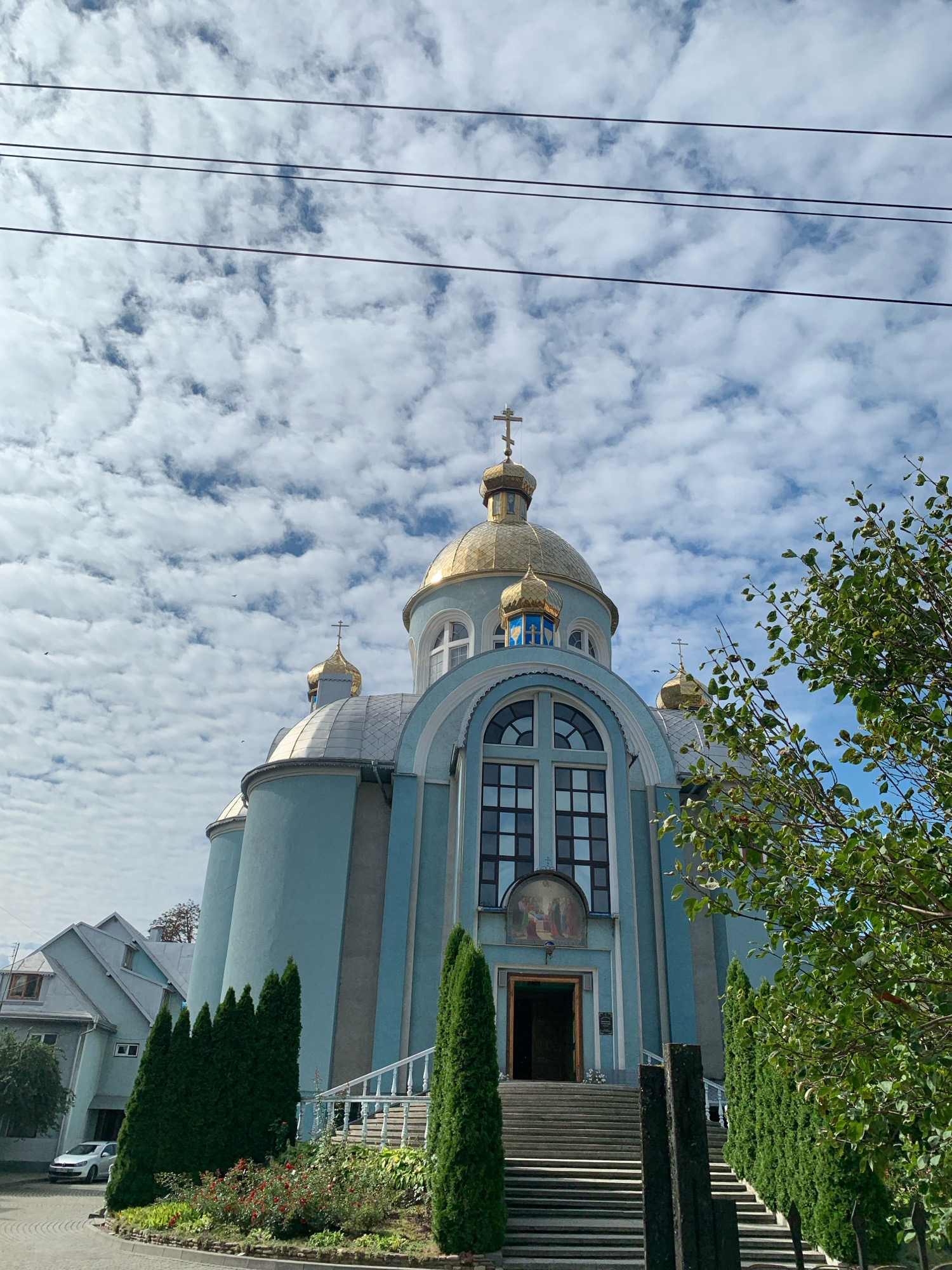 An exterior, ground-level, eye-level, low-angle shot captures a blue church with golden domes against a sky filled with scattered white clouds. The church features a grand staircase leading to its entrance, flanked by green cypress trees. To the left of the church, a neighboring light blue building with a grey roof is partially visible, with a white car parked in front of it. A tree with green leaves partially obscures the right side of the church. The image is framed by power lines crossing the top of the frame. This is the Church of the Holy Resurrection in Kolomyia, Ukraine.