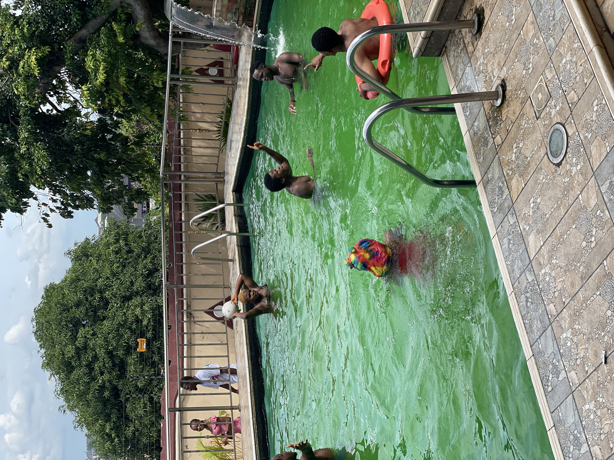 The image shows a group of people enjoying themselves in a bright green swimming pool. The pool is surrounded by decorative tilework and a metal fence. In the background, lush green trees and buildings suggest an urban or suburban setting. Several individuals are in the water, with some interacting and others engaging in solitary activities. One person is holding a white ball, another is wearing a colorful wig, and one person is seated on an orange inflatable ring. The lighting indicates it is daytime, with a partly cloudy sky.