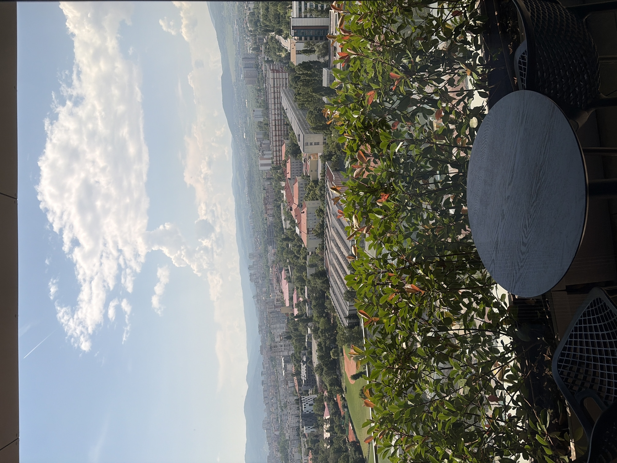 A high-angle, full shot captures a cityscape beneath a partly cloudy sky. In the foreground, a round, dark wooden table sits on a balcony, flanked by two black chairs with patterned seats. A dense hedge of green and reddish-brown leaves forms a natural barrier behind the table and chairs. Beyond the balcony, a sprawling city unfolds, with numerous buildings of varying heights and architectural styles dotting the landscape. Distant mountains form a hazy backdrop under a pale blue sky with scattered white clouds. A contrail from an aircraft is visible in the upper left corner of the sky. The overall scene suggests a serene observation point overlooking an urban environment.
