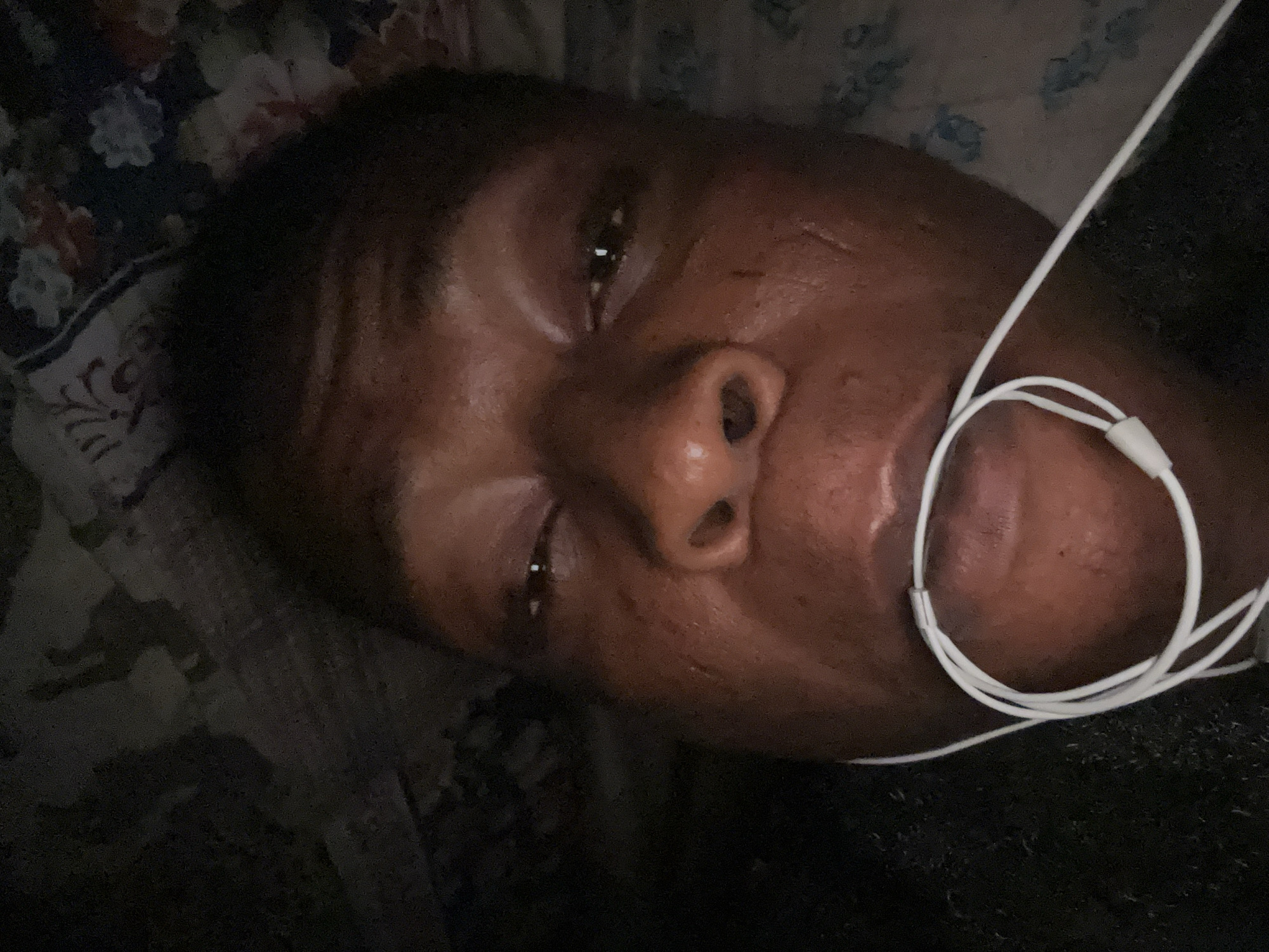 A close-up image shows a dark-skinned person lying on a floral-patterned surface, possibly a bed or cushion. The person has earbuds positioned around their neck. The location is unknown.