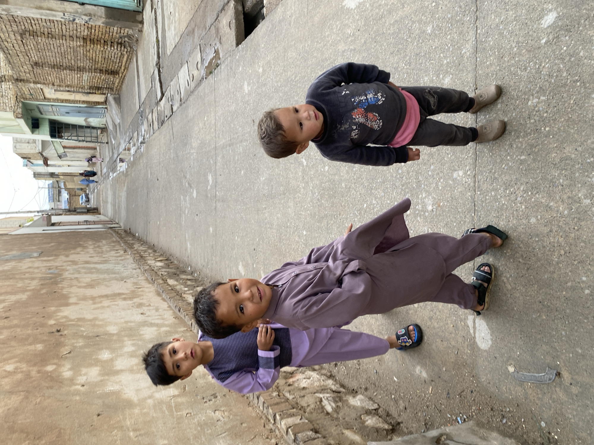 This image depicts an urban alleyway in Kandahar, Afghanistan, under an overcast sky. In the foreground, three young boys are standing on a concrete path.
The boy on the left, estimated to be 6-8 years old, wears a purple shalwar kameez with a horizontally striped dark top and lighter purple sleeves and trousers. He wears blue and black sandals with colorful accents and looks directly at the camera with a neutral expression.
The central boy, estimated to be 5-7 years old, wears a solid medium-purple shalwar kameez and black sandals. He faces the camera with a slight smile.
The boy on the right, estimated to be 2-4 years old, wears a dark grey long-sleeved top with a faded red, blue, and white graphic design, a pink t-shirt underneath, dark grey trousers, and light brown soft-soled shoes. He looks slightly to his left with a curious expression.
The concrete path shows wear and is partially wet, with small puddles in a narrow concrete drainage channel running along the right side. A bent piece of dark metal debris is on the ground in the lower-left foreground.
To the left, a tall, light brown/beige wall, possibly mud brick or adobe, shows signs of age and texture. To the right, buildings constructed of exposed brickwork and some with painted sections line the street. Further down the alley, a white car is parked, and several adult figures, including two women in burqas, are walking away from the camera. Buildings in the distance exhibit varying construction materials and conditions, with solar panels visible on some rooftops.