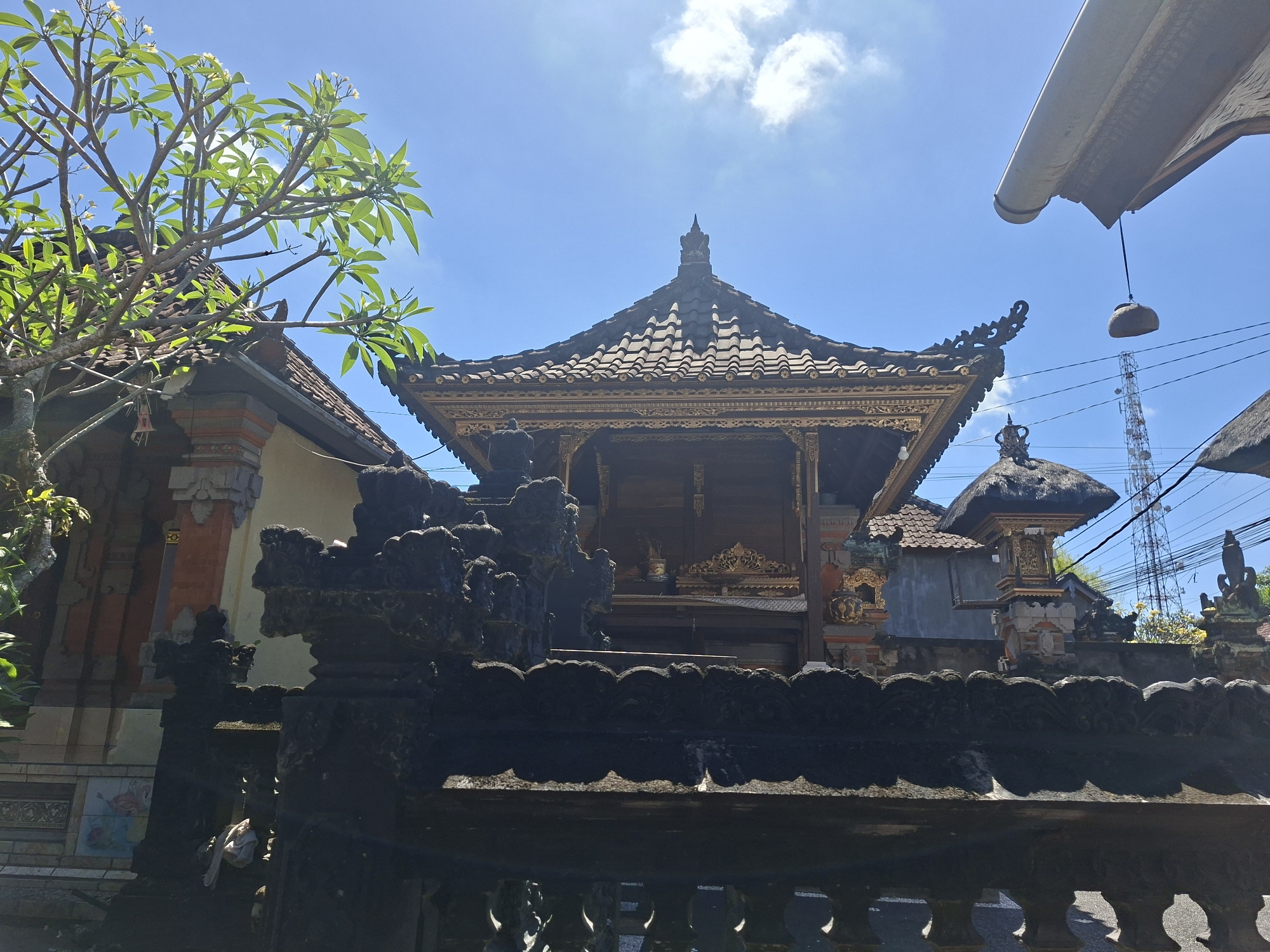 The image presents a bright, sunlit view of traditional Balinese architecture, set against a clear blue sky, consistent with the provided location context of Blahbatuh, Indonesia.
In the center of the frame stands a prominent Balinese temple or pavilion. It features a dark, multi-tiered tiled roof crowned with an ornate finial. Beneath the roof, the structure is adorned with intricate golden carvings and decorative elements, showcasing detailed craftsmanship.
The immediate foreground is dominated by a dark, elaborately carved stone fence or wall, which stretches across the bottom of the image and partially obscures the view.
To the left, a lush green plumeria (frangipani) tree with small white blossoms extends into the frame, its leaves brightly lit by the sun. Behind the tree, another building with a tiled roof and a patterned wall can be seen.
On the right side, a smaller, traditional Balinese shrine or bale with a thatched roof is visible. Further in the background, a tall metal communication tower rises, accompanied by overhead power lines. A white awning or roof overhang, with a rock-like object suspended by a string, is partially visible in the top right corner.
The time of day is clearly bright and sunny, indicated by the brilliant blue sky and strong shadows. A few scattered white clouds are present. No activity or specific event is depicted; the scene is static. There is no visible text in the image.
