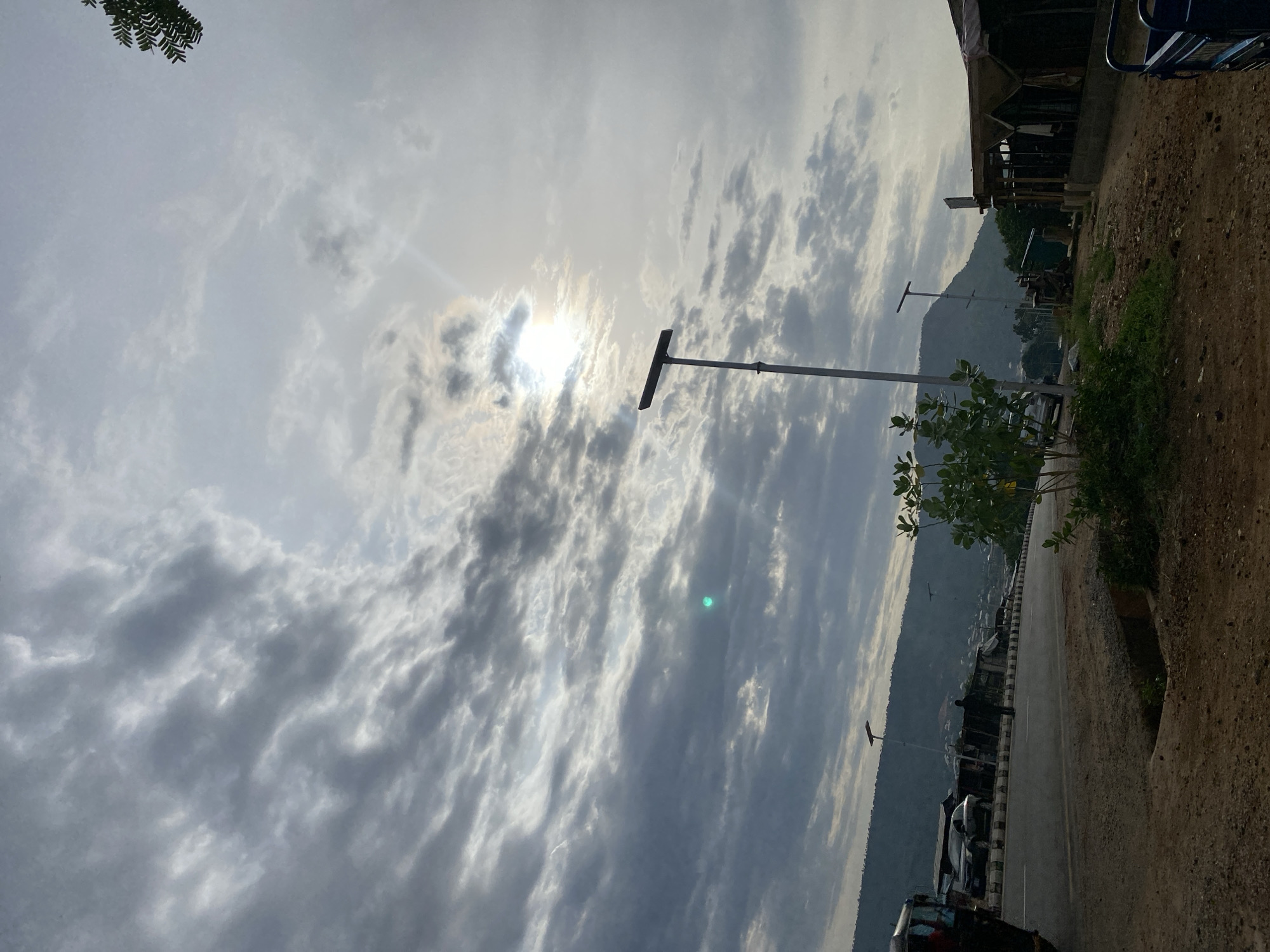 The image depicts a road in Adankolo, Nigeria, with a mountain range in the background. The sky is overcast with the sun obscured by clouds. A utility pole stands along the roadside. Vegetation grows along the edge of the road. A building is visible on the right side of the road. Several vehicles are parked along the opposite side of the road. A patch of trees in the foreground is on the right side of the photo.