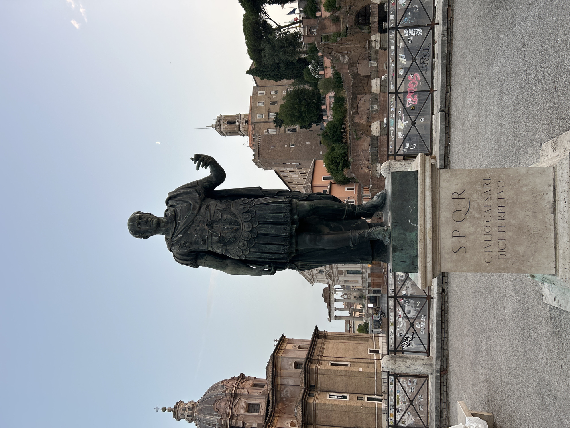 A full-body bronze statue of a man in Roman military attire stands on a stone pedestal in the foreground. The statue is positioned in the center of the frame, facing slightly left and with his right arm extended and index finger pointing upwards. The pedestal has an inscription in Latin that reads "SPQR, CIVLIO CAESARI, DICT PERPETVO". Behind the statue, a fenced-off area with graffiti-covered metal bars separates the viewer from ancient Roman ruins and historic buildings. To the left, a large dome structure with a cross on top dominates the skyline. Further back, a tower with a pointed roof rises above a multi-story building. The sky is clear and a crescent moon is visible. The overall scene appears to be set in Rome, Italy, likely at the Roman Forum, given the presence of ancient ruins and iconic Roman architecture.