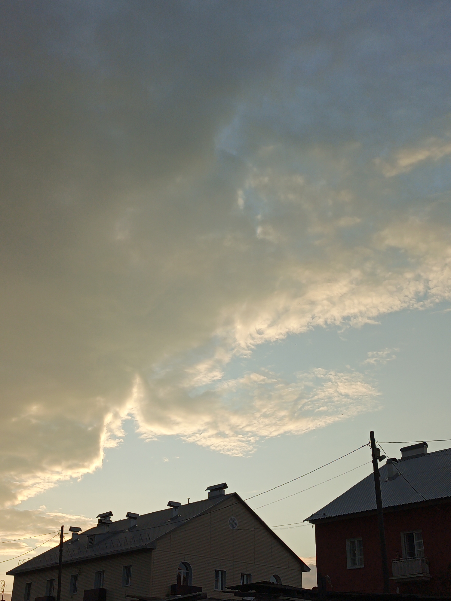 The image captures a view of buildings against a cloudy sky, likely in Medvedyovka, Russia. The sky exhibits a dramatic contrast between dark, heavy clouds on the left, tinged with yellow from what appears to be sunset or sunrise light, and lighter, wispy clouds on the right. Two buildings are visible in the foreground. The building on the left is lighter in color with multiple dormer windows on its sloped roof. The building on the right is red with a dark roof, featuring a chimney and windows, and a small balcony. Power lines stretch between utility poles, crossing the scene in front of the buildings and into the sky. The overall atmosphere is calm, with no people or specific actions depicted.