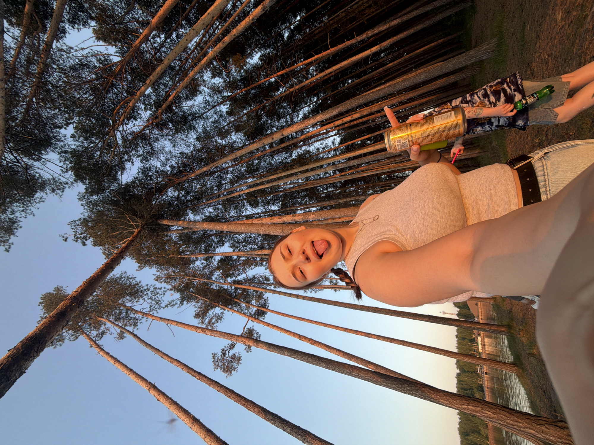 The image shows a selfie taken from a low angle. A young woman with a braid is holding a can of drink, sticking her tongue out at the camera. Behind her are tall pine trees reaching towards the sky, and in the far background, a body of water with trees on the opposite bank. Another person holding a green glass bottle can partially be seen. Based on the landscape and composition, this scene may be in the city of Chaikovsky, Russia, or a similar environment with pine forests and lakes.