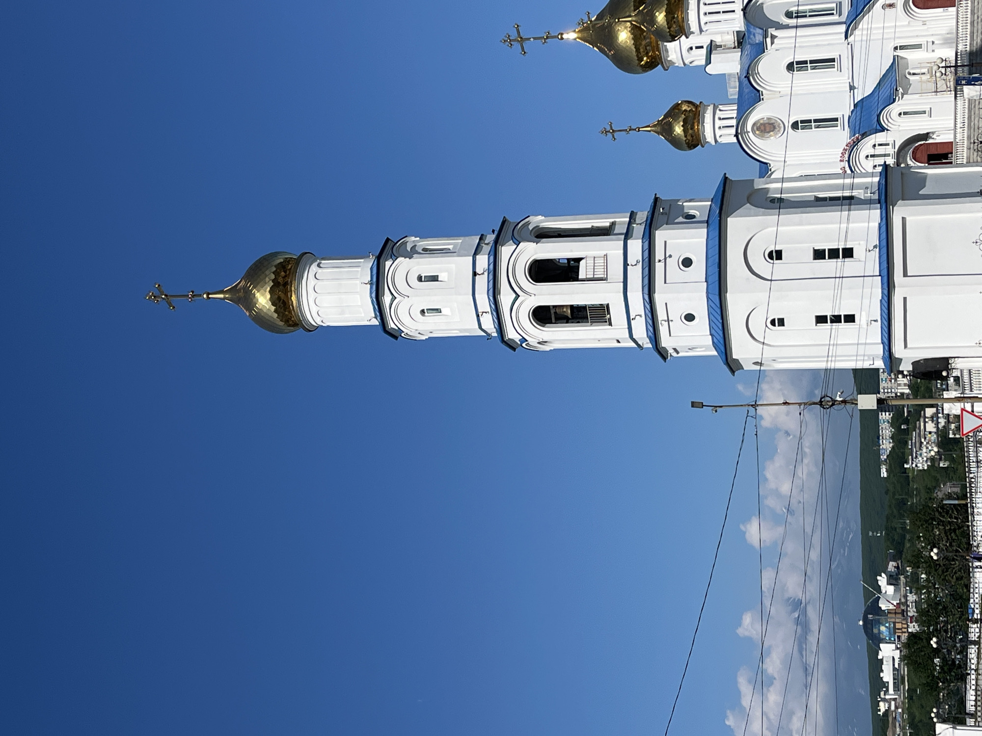 An upward angle shot captures the bell tower and domes of the Holy Trinity Cathedral against a clear blue sky in Petropavlovsk-Kamchatsky, Russia. The white cathedral features striking blue roofs and golden domes topped with crosses. The bell tower, also white with blue trim, has arched openings with a bell visible in one. Below the cathedral, a cityscape with various buildings, including one with a distinctive dome, is visible amidst green trees, under a sky dotted with white clouds. Power lines crisscross the foreground.