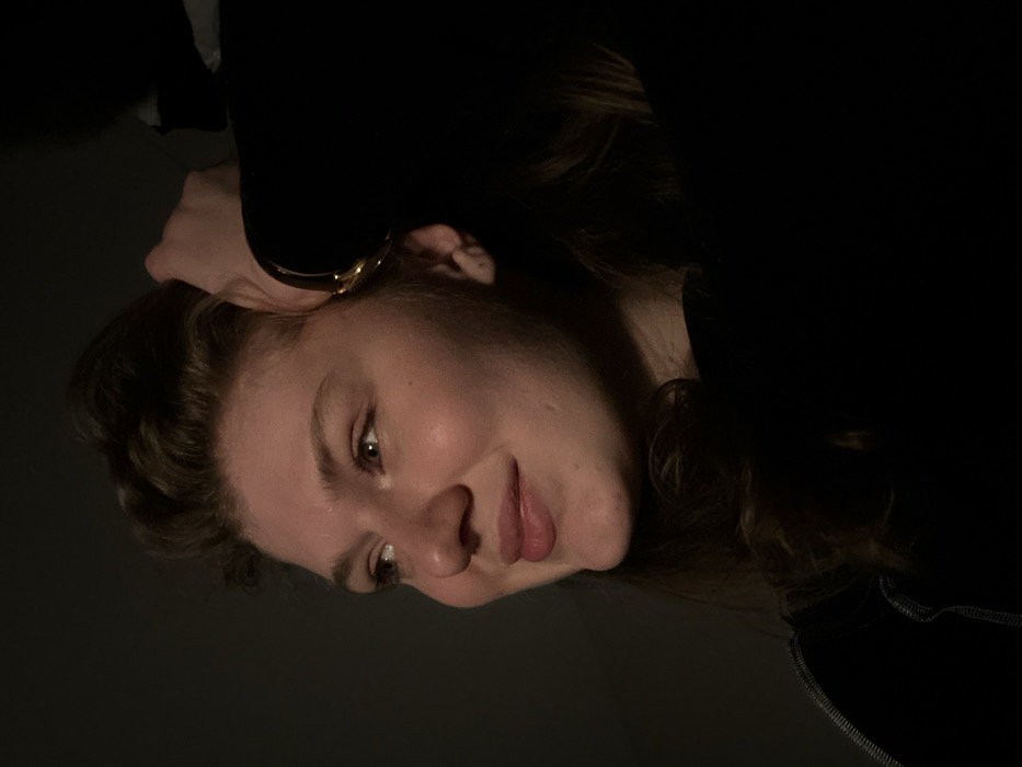 A close-up portrait depicts a young adult female, estimated between 20-30 years of age, with light skin, hazel eyes, and light brown wavy hair. She is positioned in profile, facing left, with her head slightly tilted and a subtle smile on her lips, gazing to the left. A distinct light source illuminates the left side of her face, highlighting her cheek, nose, and forehead, while the right side of her face and much of the scene are cast in deep shadow. Her right hand, wearing a gold-colored bracelet on the wrist, is raised and appears to be touching or adjusting her hair near the top right of her head. She is clad in a dark garment, possibly a high-neck top, with faint light-colored stitching visible on the lower left portion of the frame, near her shoulder. The background is uniformly dark and offers no discernible environmental details. The described location for this image is Kazan, Russia.