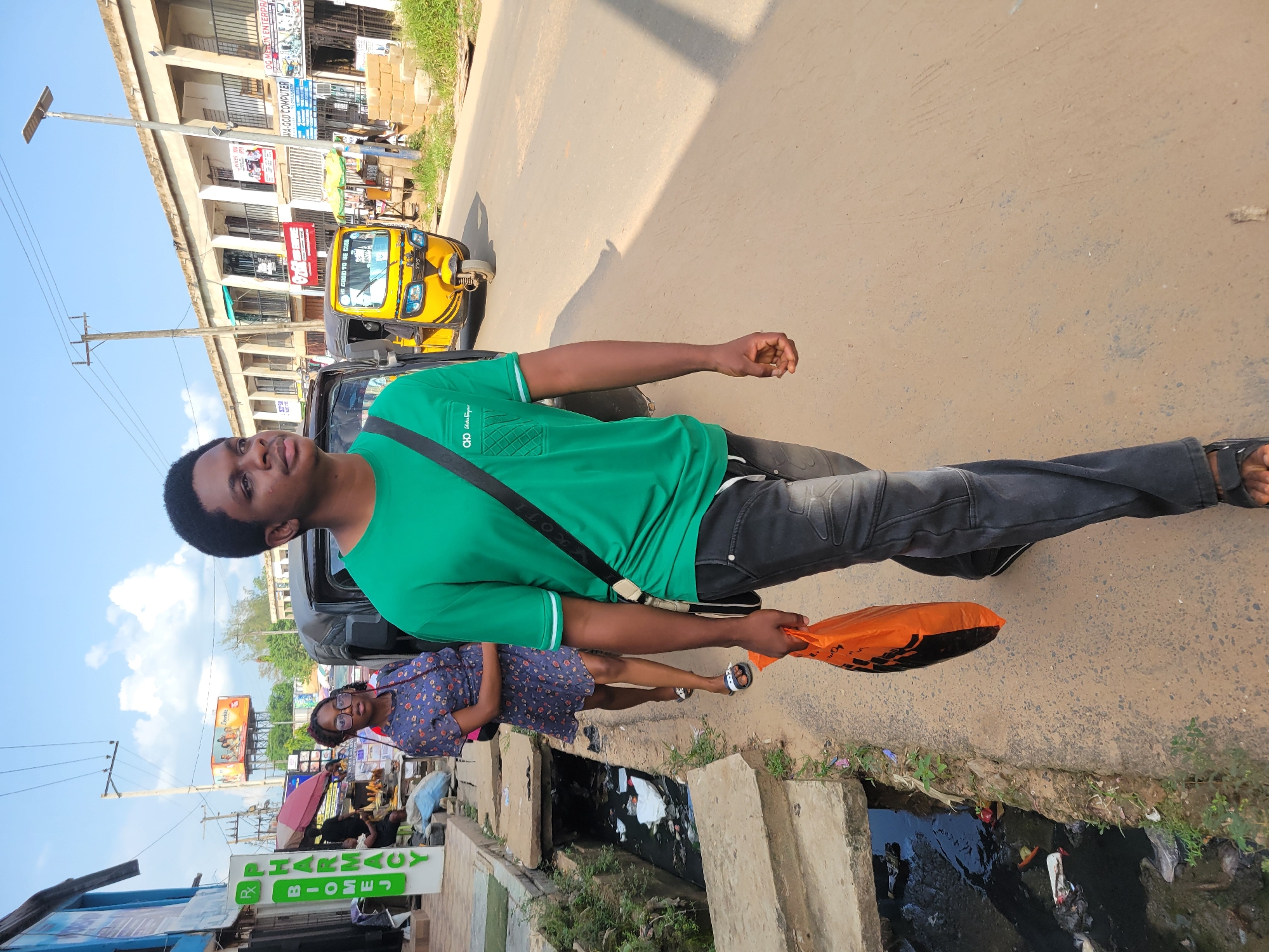 The image shows a young man in a green t-shirt and dark jeans standing in an outdoor urban environment. He is holding an orange bag in his right hand and has a black strap across his chest. Behind him, a woman in a patterned dress is visible, and further in the background, a yellow auto-rickshaw is parked. The scene appears to be a street with buildings lining one side and a gutter filled with trash on the other.
Visible text includes "PHARMACY" and "BIOMEJ" on a green sign, "GOOD TO BE COON" on the auto-rickshaw, and "OG MERLIN ENTERPRI" and "WA-GOD COMPUTER" on signs on the buildings. The sky is blue with scattered clouds, suggesting it is daytime. The overall setting seems to be a busy commercial area in Egbagu, Nigeria.
