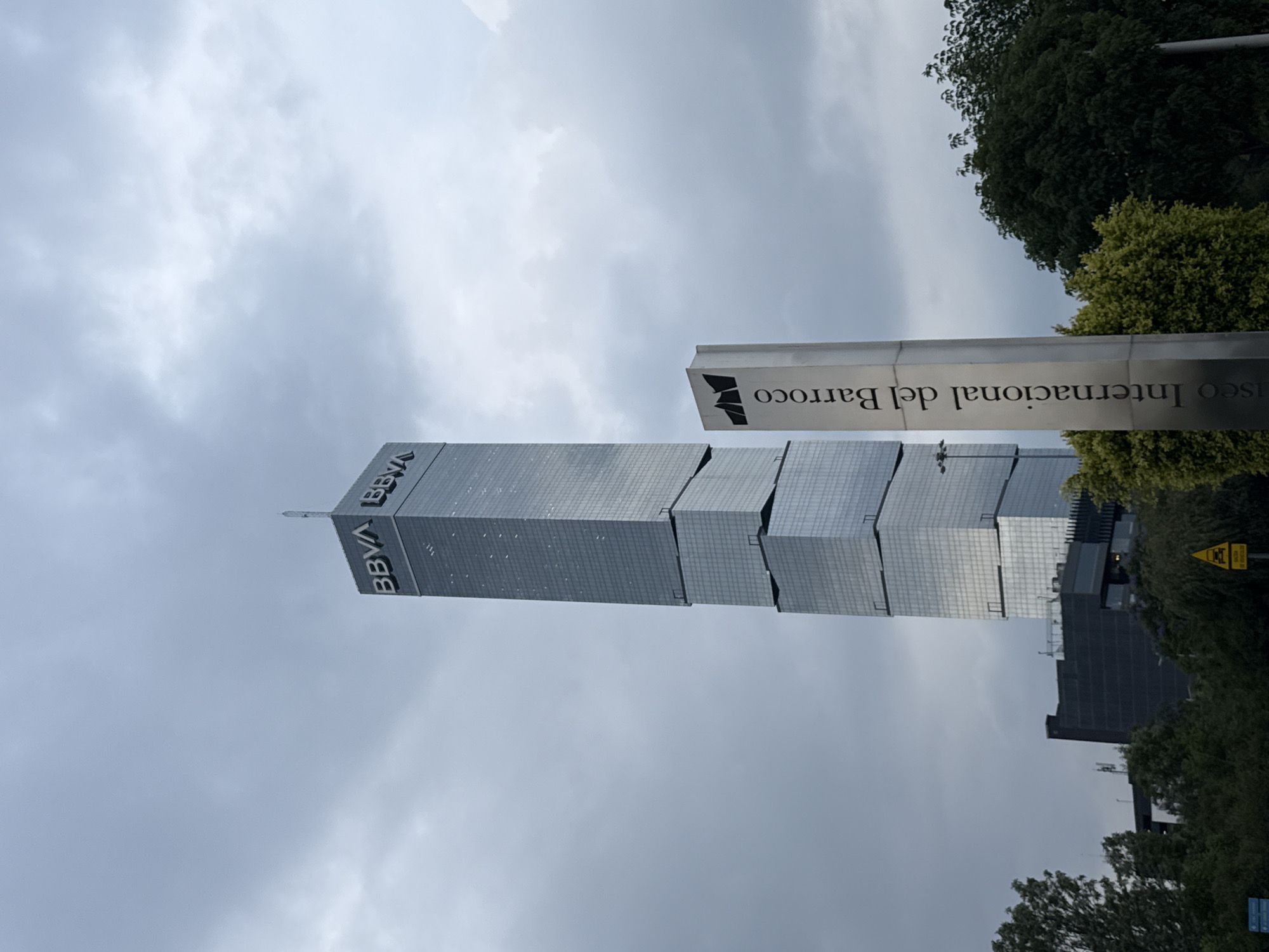 The image shows a low-angle view of the BBVA Tower in Puebla, Mexico. The tower is a modern skyscraper with a stepped design, constructed from reflective materials. The sky is overcast. In the foreground, a sign reads "Museo Internacional del Barroco". Trees surround the tower.